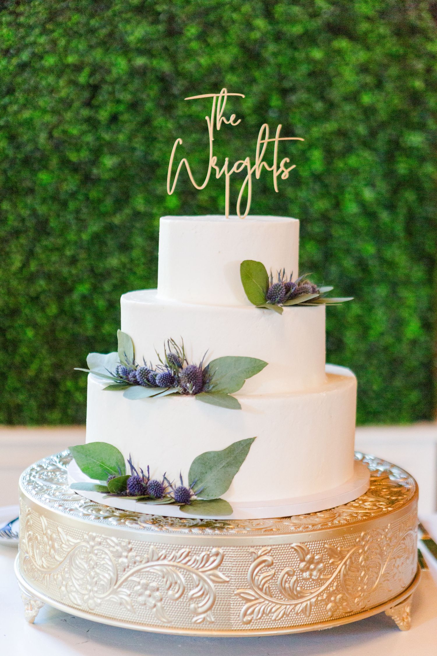 Three-tier white wedding cake with greenery and purple flowers on silver cake stand.