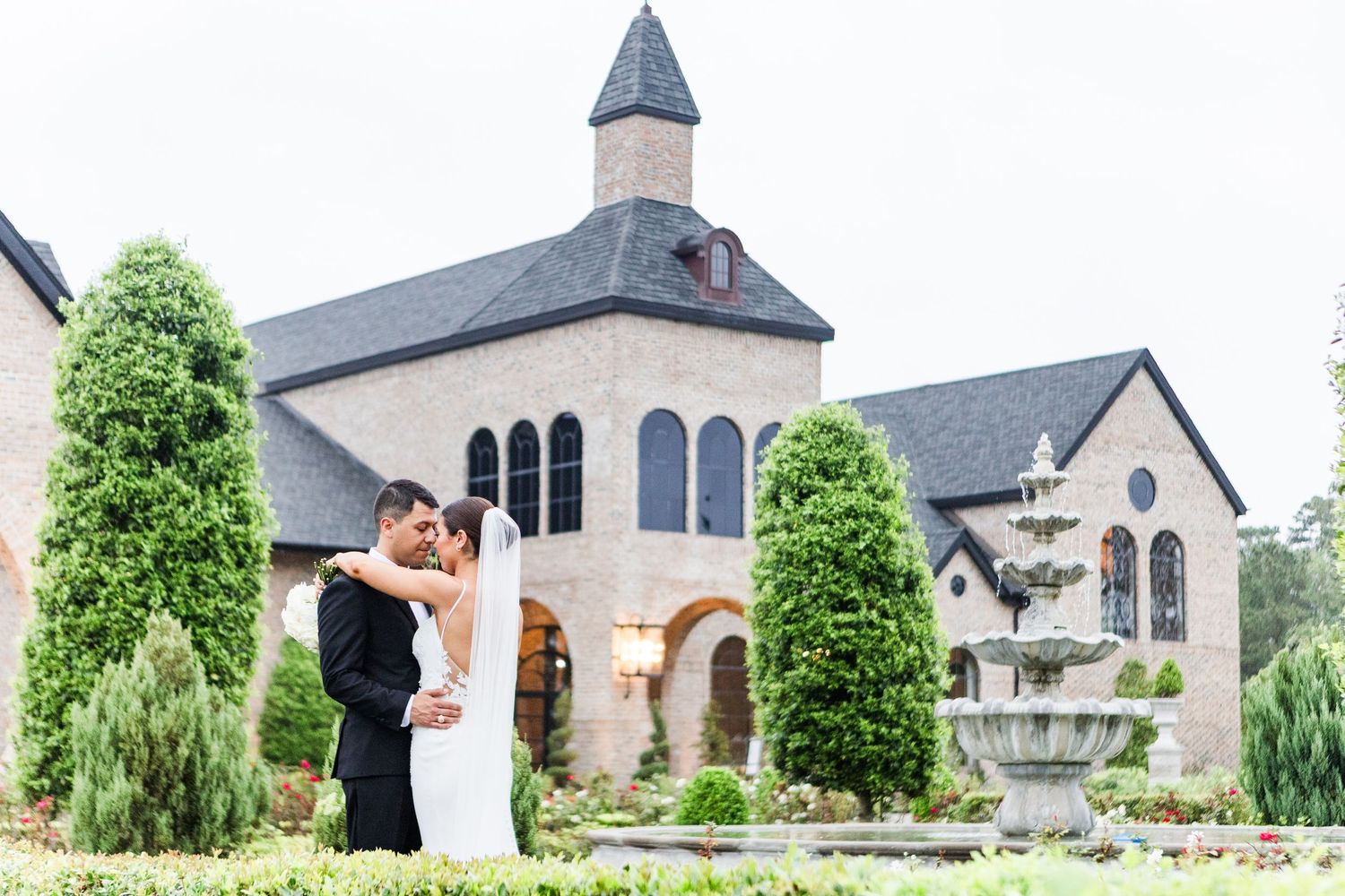 Couple embracing in front of a beautiful chapel building with fountain and landscaped gardens.