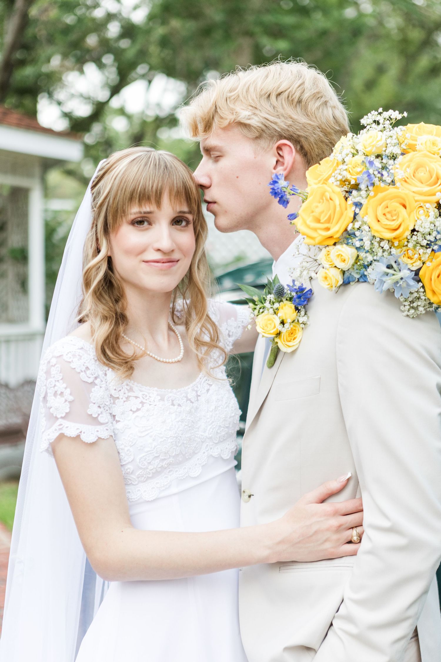 Wedding couple with yellow and blue floral bouquet against outdoor backdrop.