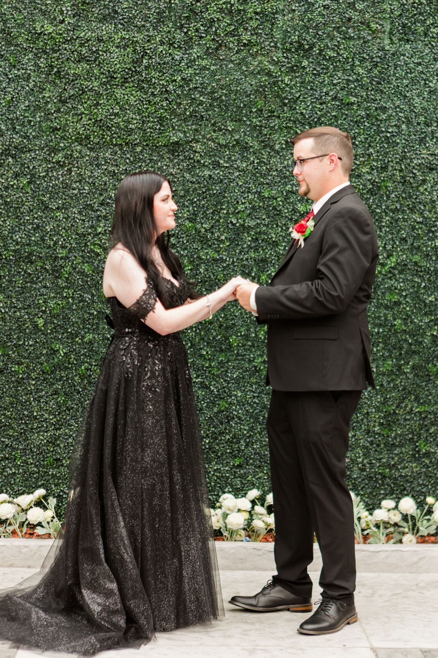Couple hold hands during wedding ceremony against green hedge background.