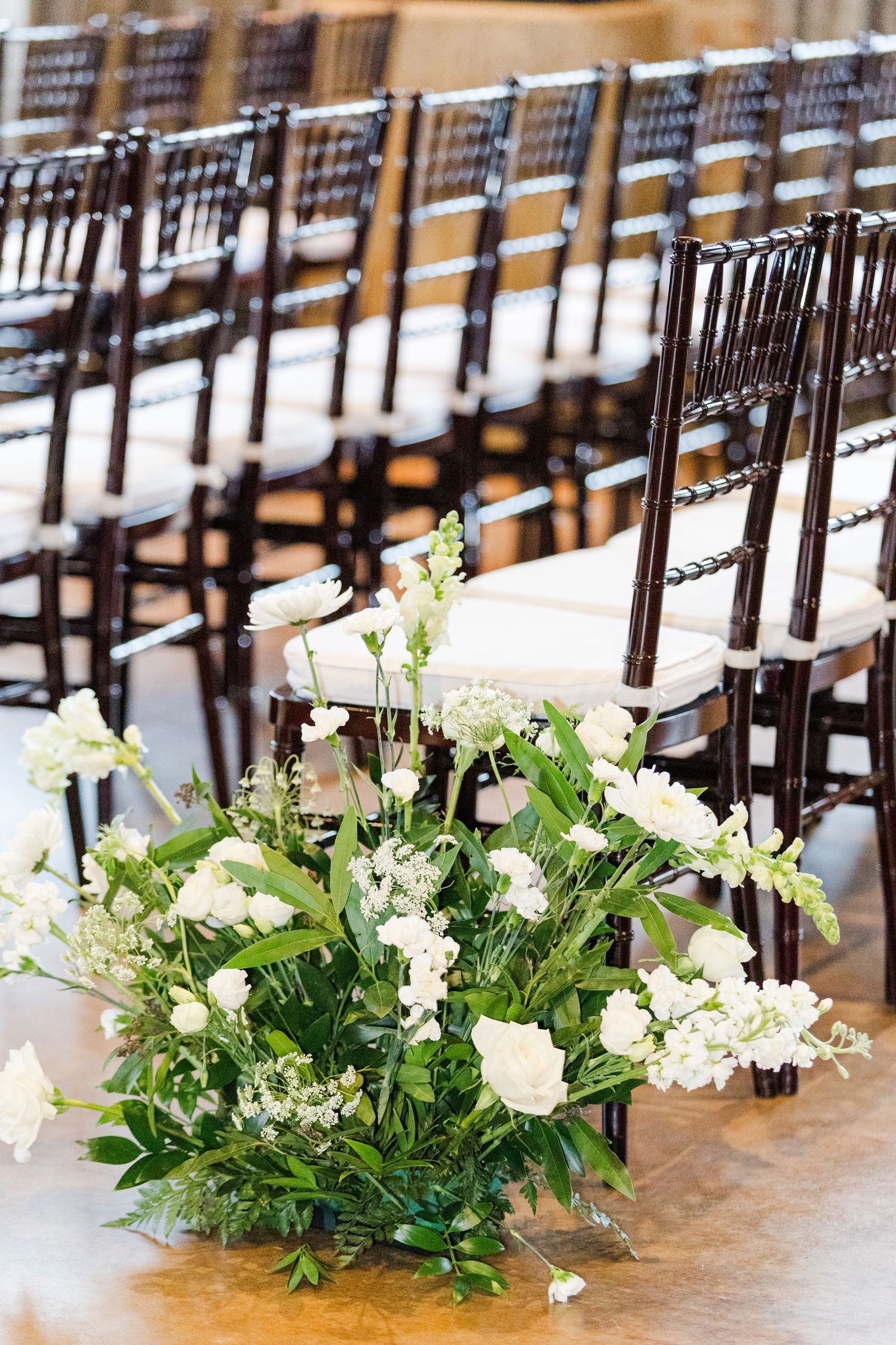 Rows of dark wooden chairs line an indoor ceremony space with elegant white floral aisle decorations.