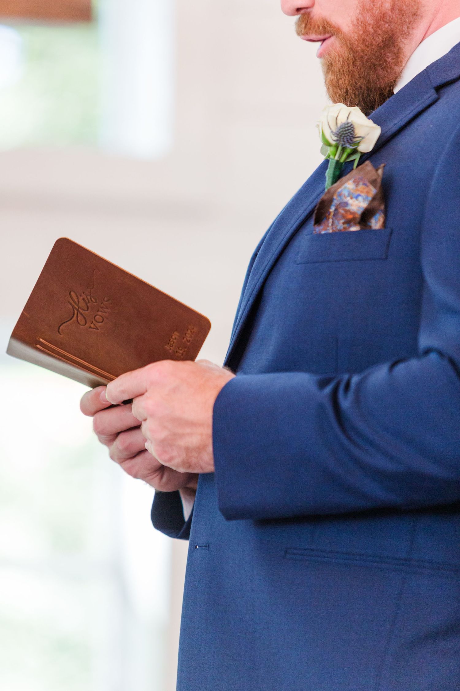 Close up of groom in navy suit holding brown leather book with boutonnière.
