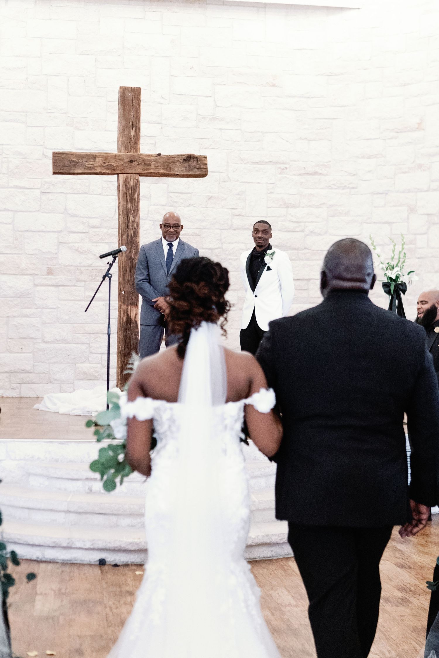 A wedding ceremony takes place in a minimalist white chapel with a wooden cross on the wall.