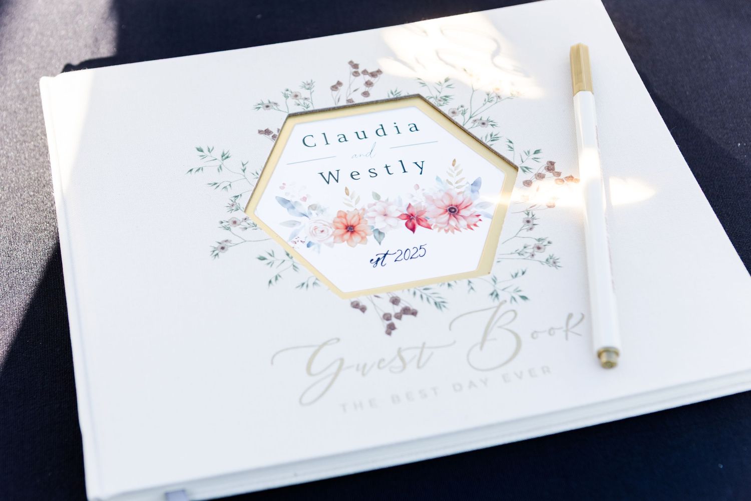 White wedding guest book with gold hexagonal design and delicate floral watercolor details, displayed with a gold pen.