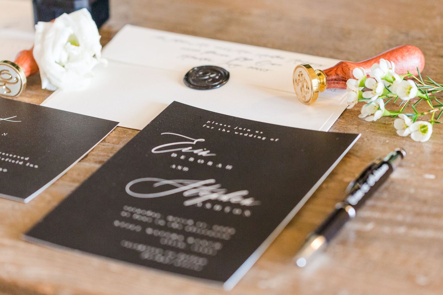 Black wedding invitations with silver calligraphy laid out on rustic wooden surface.