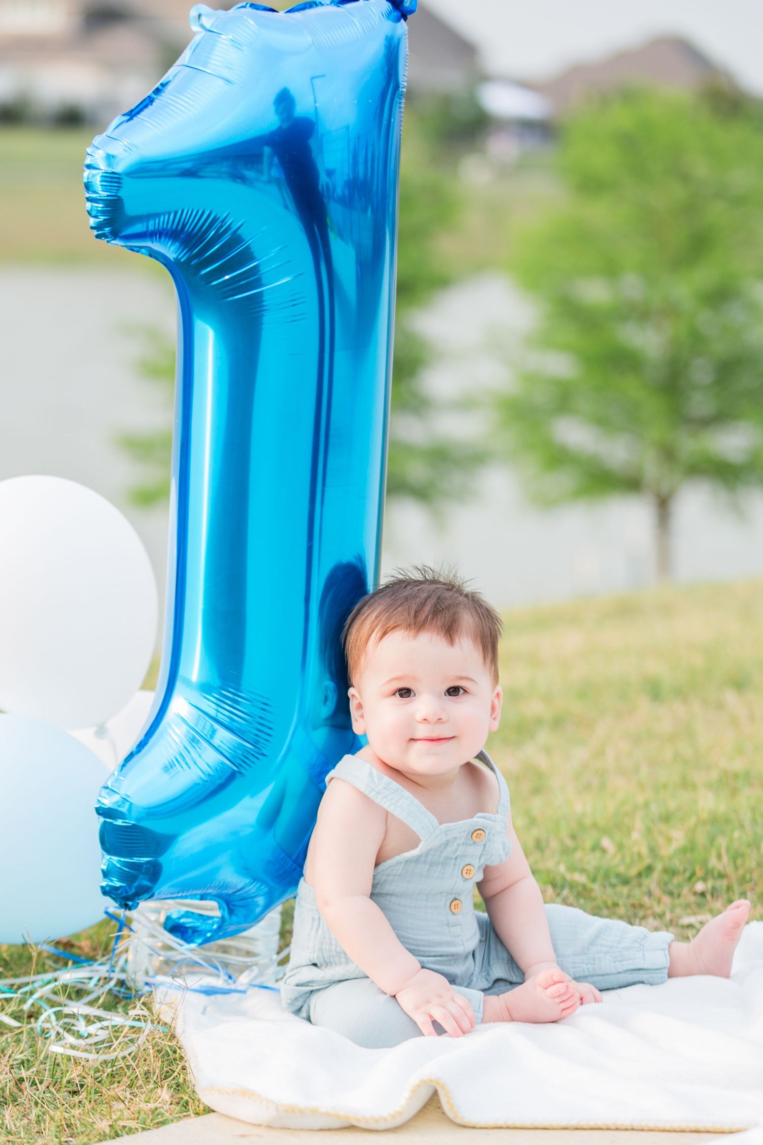 Baby in gray outfit sits next to large blue number one balloon on grass.