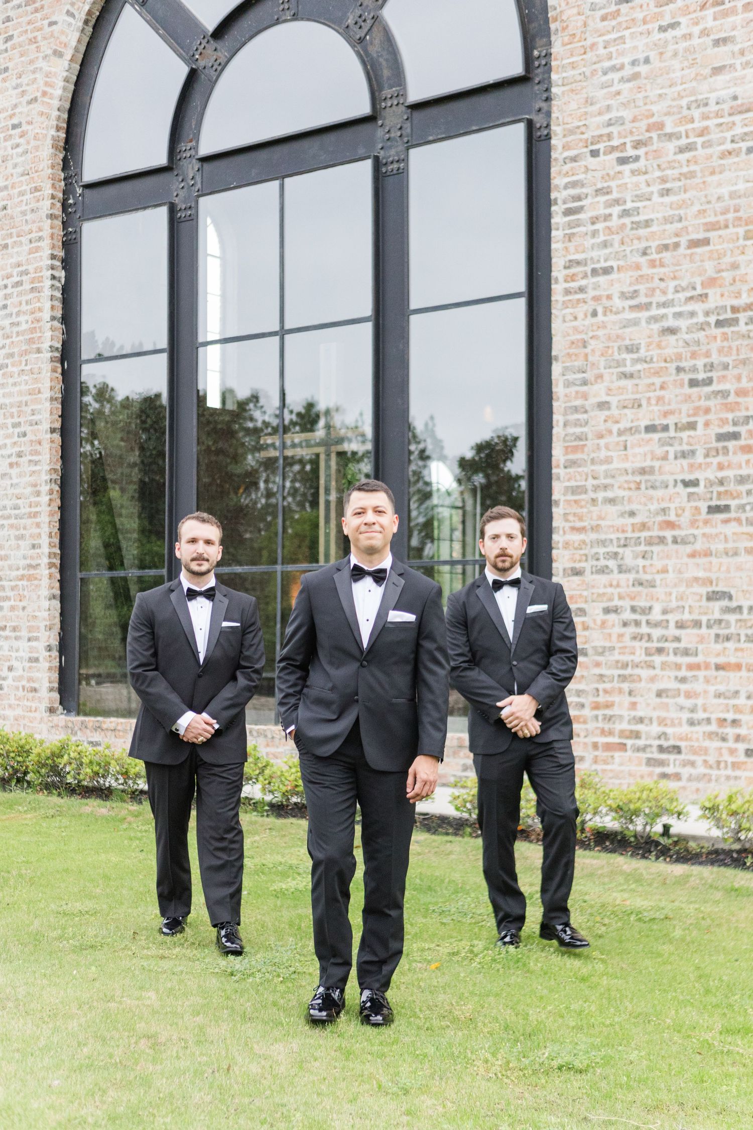 Wedding party in black tuxedos walking and posing outside brick building with arched windows.