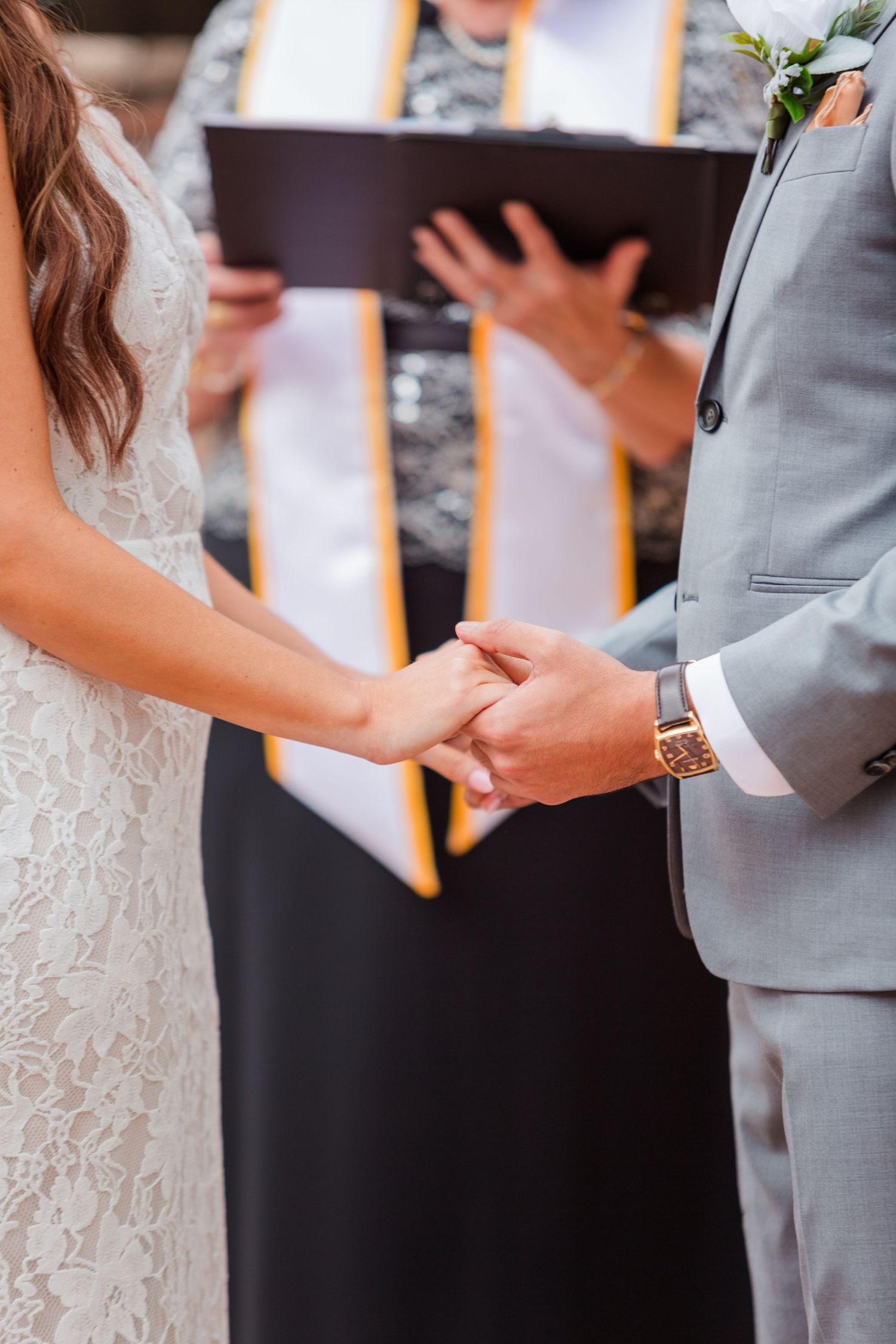 Close-up of hands during a wedding ceremony exchange with white lace dress and gray suit visible.