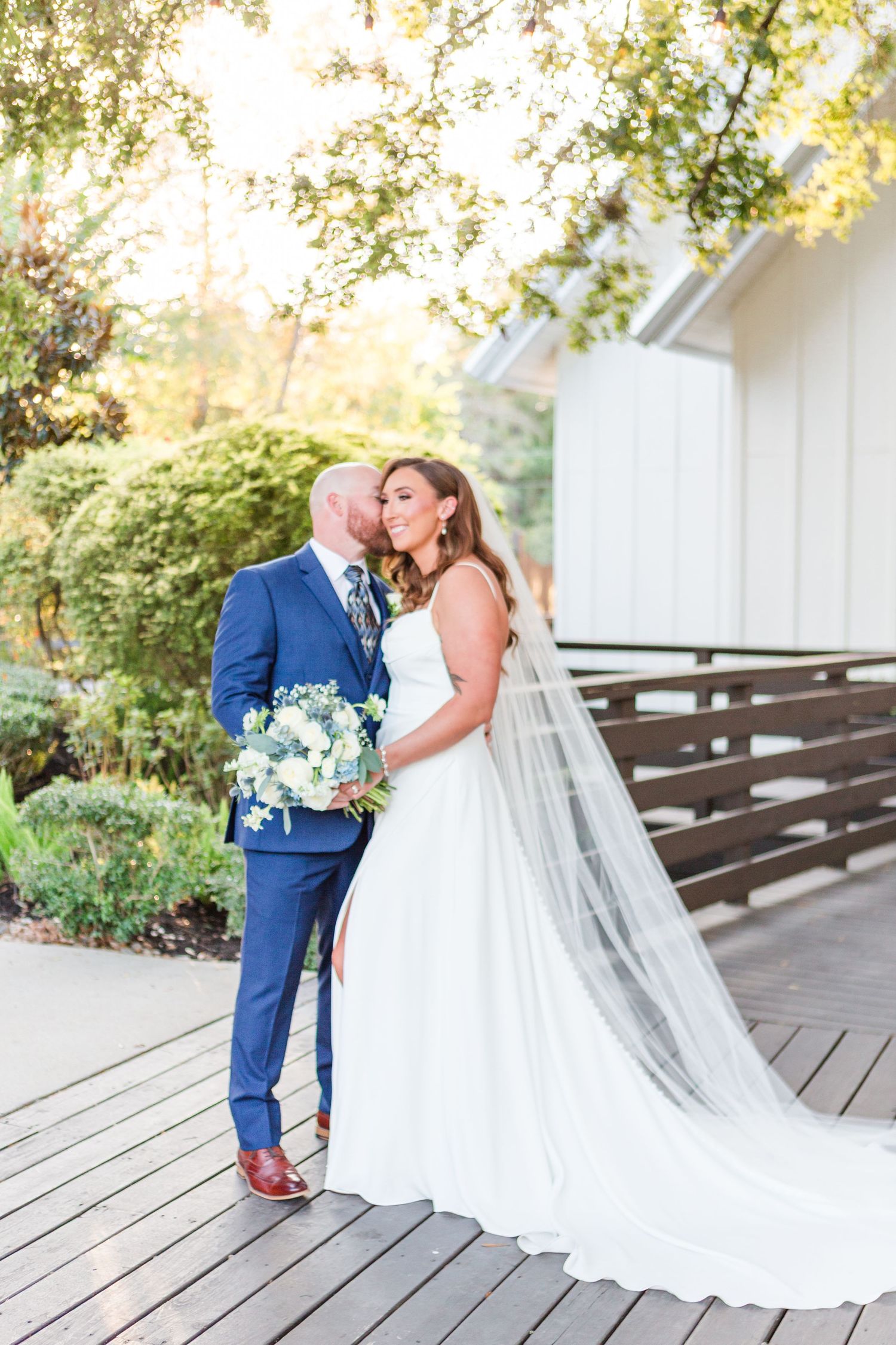 Romantic wedding couple shares intimate moments on wooden deck surrounded by lush greenery during summer photo session.