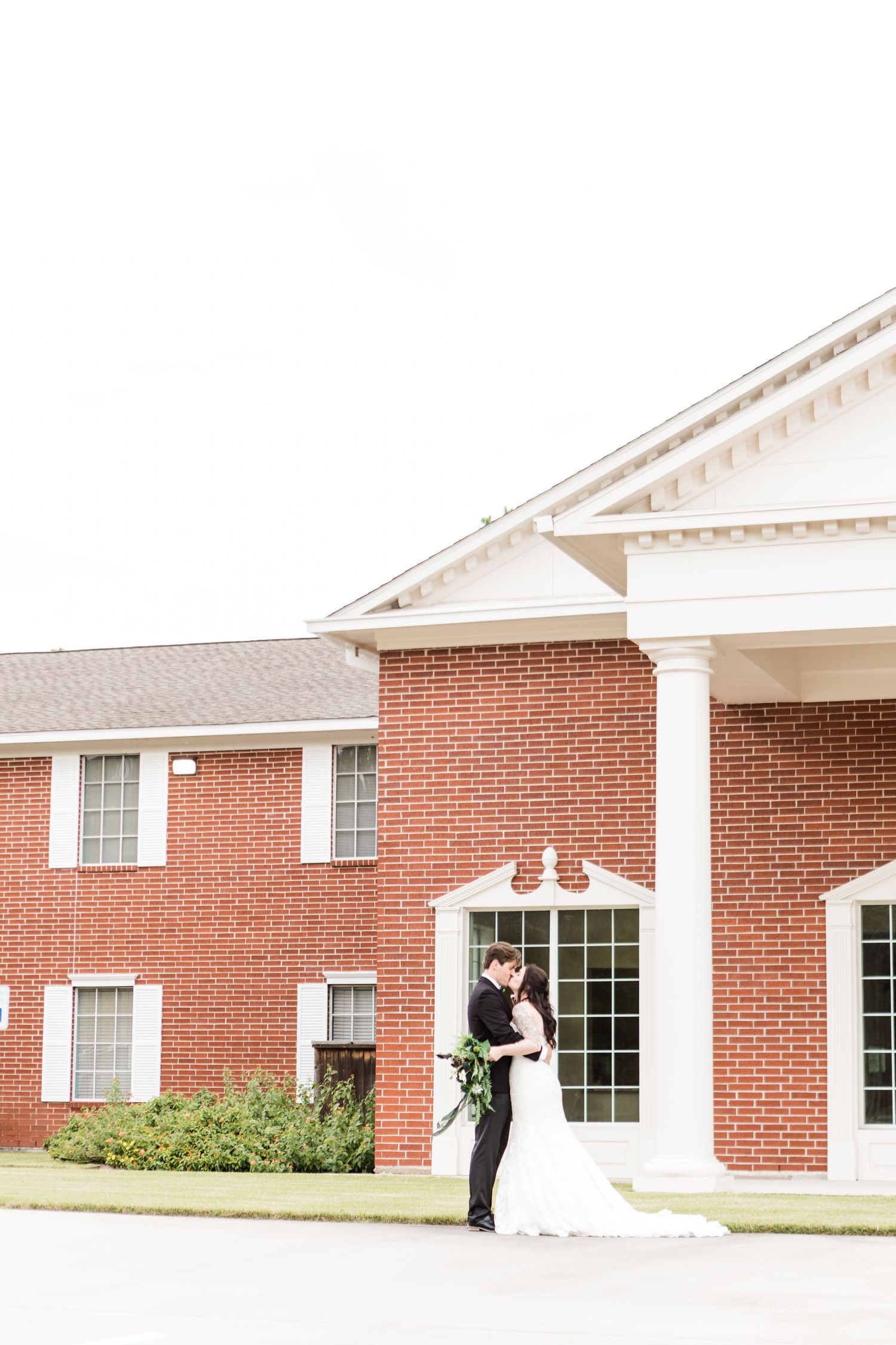 Wedding couple shares romantic moments outside brick building with white architectural details.