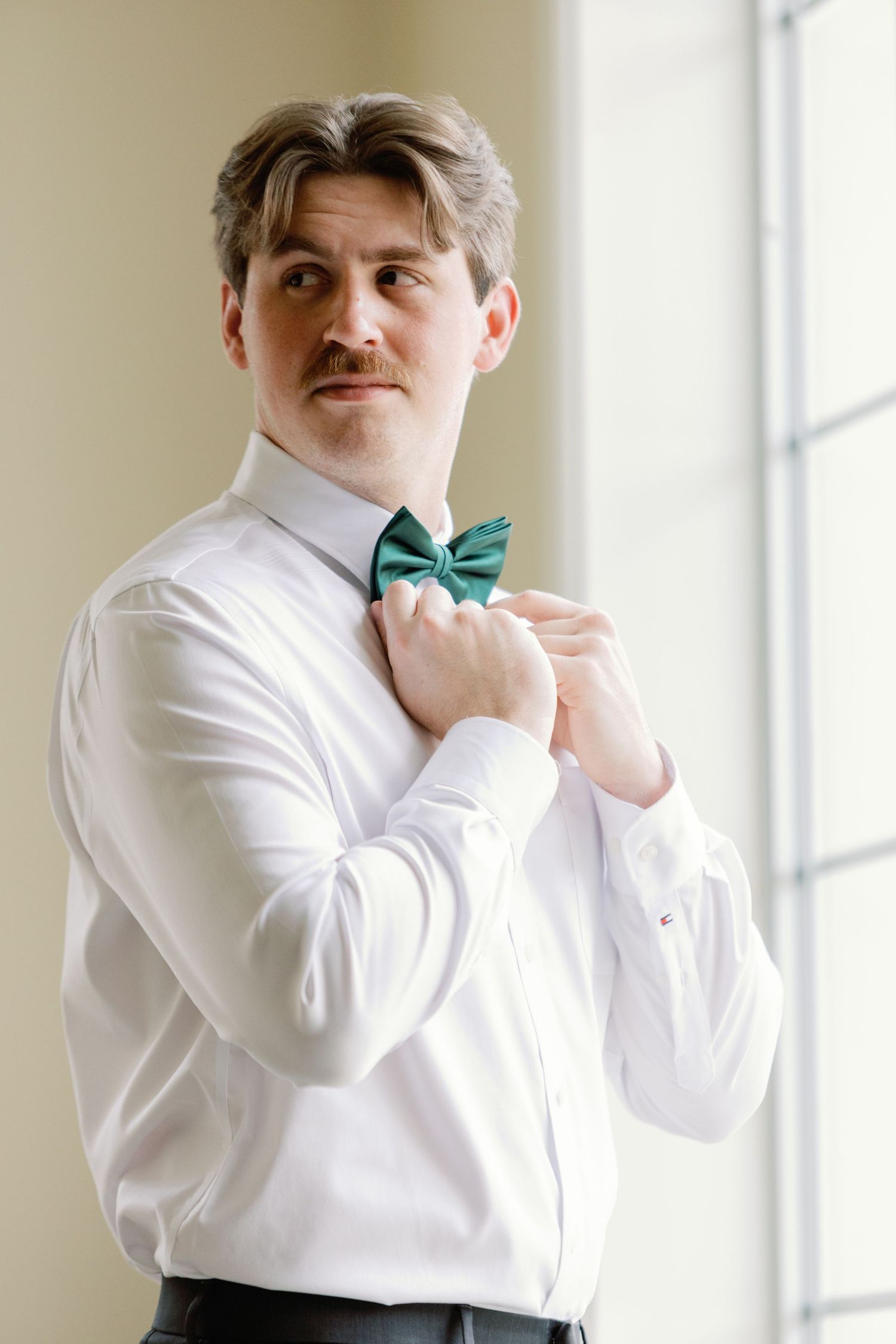 Person adjusts emerald green bow tie while wearing crisp white dress shirt in sunlit room.