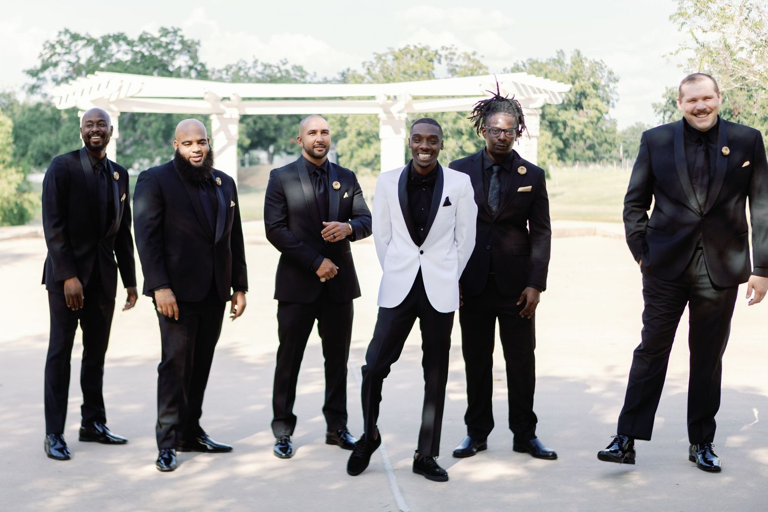 Groomsmen in black suits and one in white pose outdoors for a wedding photo.