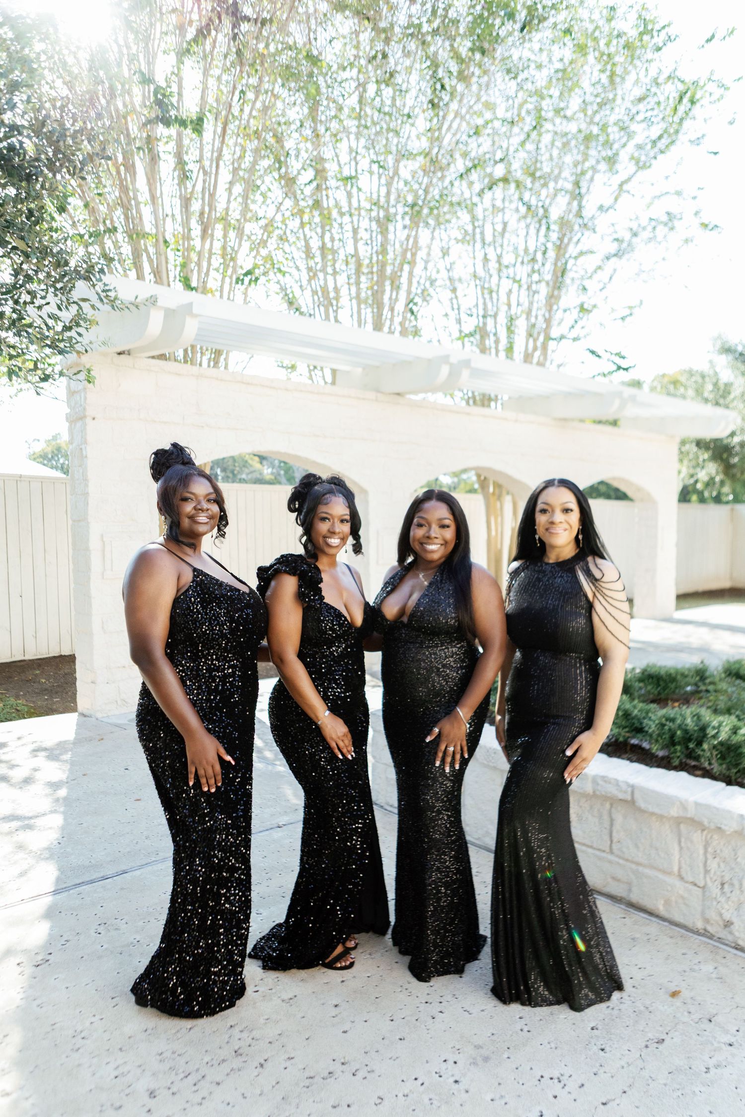Wedding party in elegant black formal dresses poses outdoors by a white stone wall.