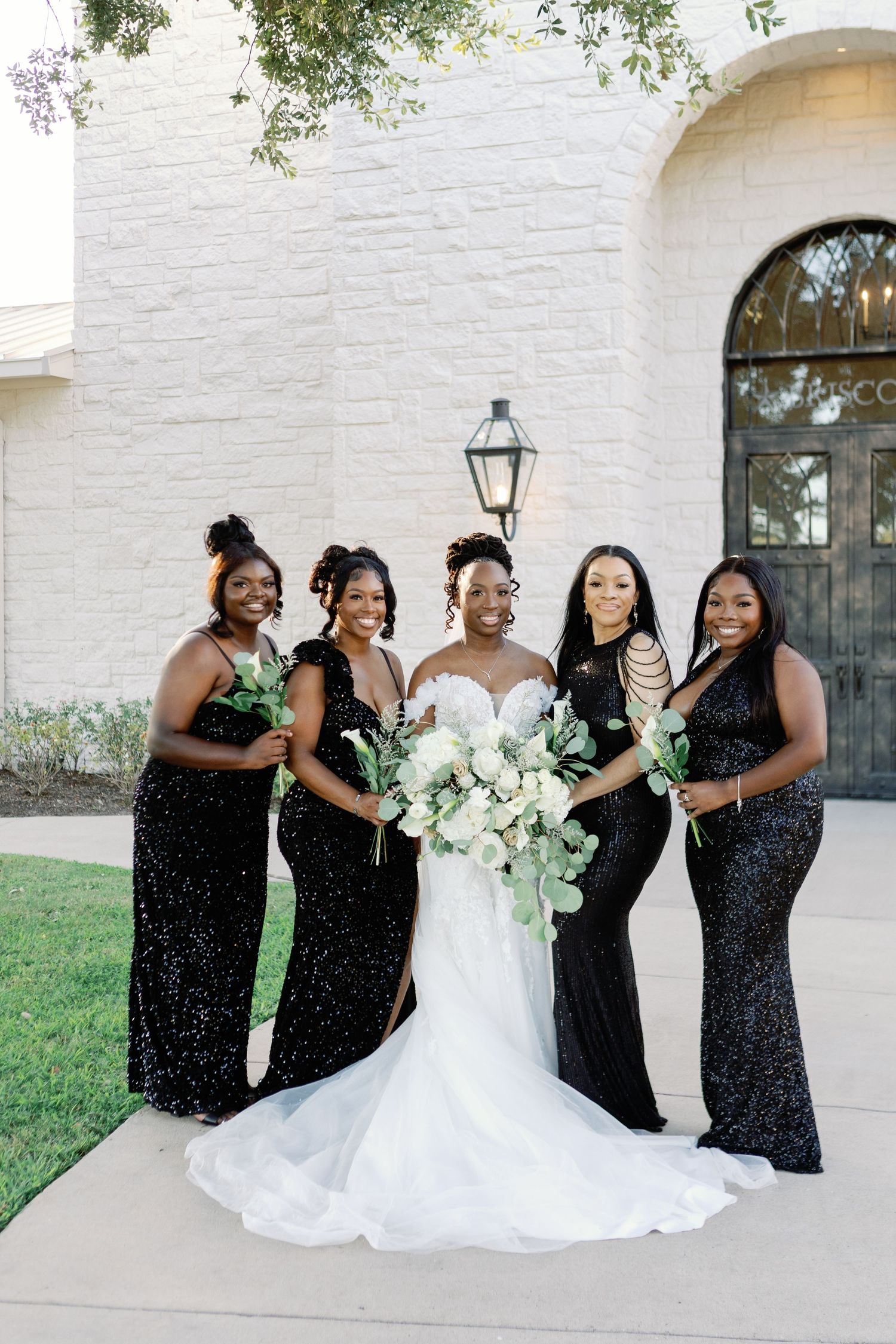 A bridal party in black sequined dresses poses together outside a white building with arched windows.
