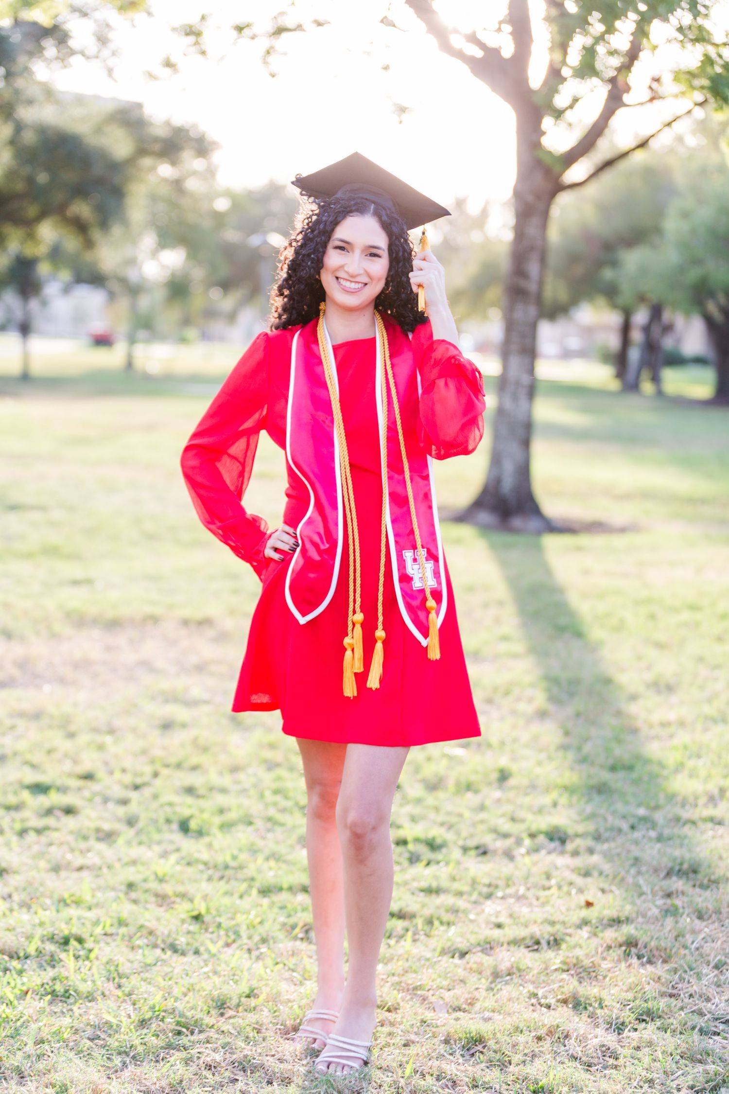 Smiling graduate in red dress and cap with honor cords poses outdoors in sunny park setting.