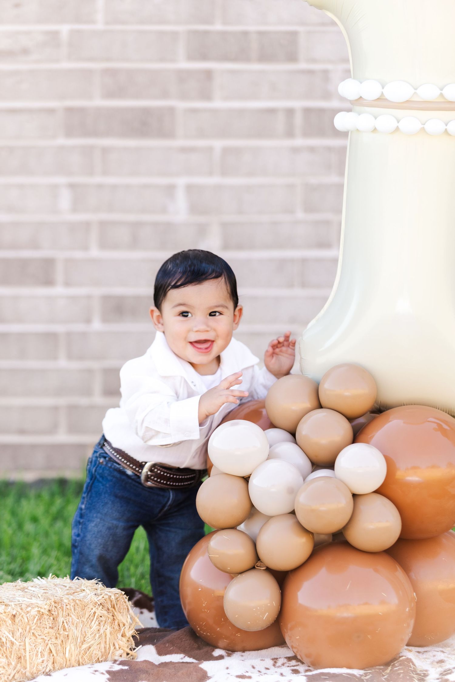 Child smiles while playing near brown balloons and hay bales against brick wall.