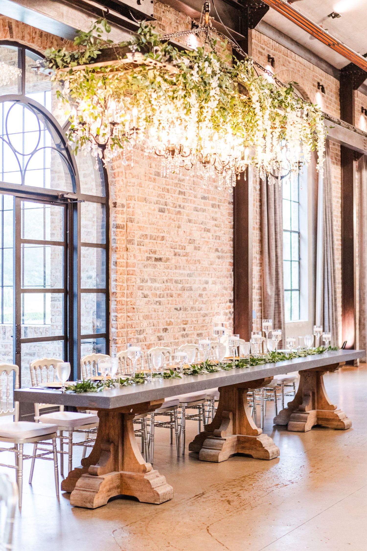 Elegant wedding venue with exposed brick walls, large windows, and wooden tables draped in white linens and floral decor.