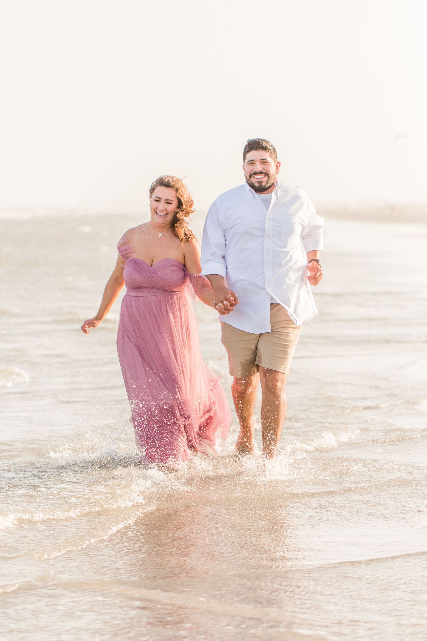 A couple splashes and plays in ocean waves during a beach photoshoot at sunset.