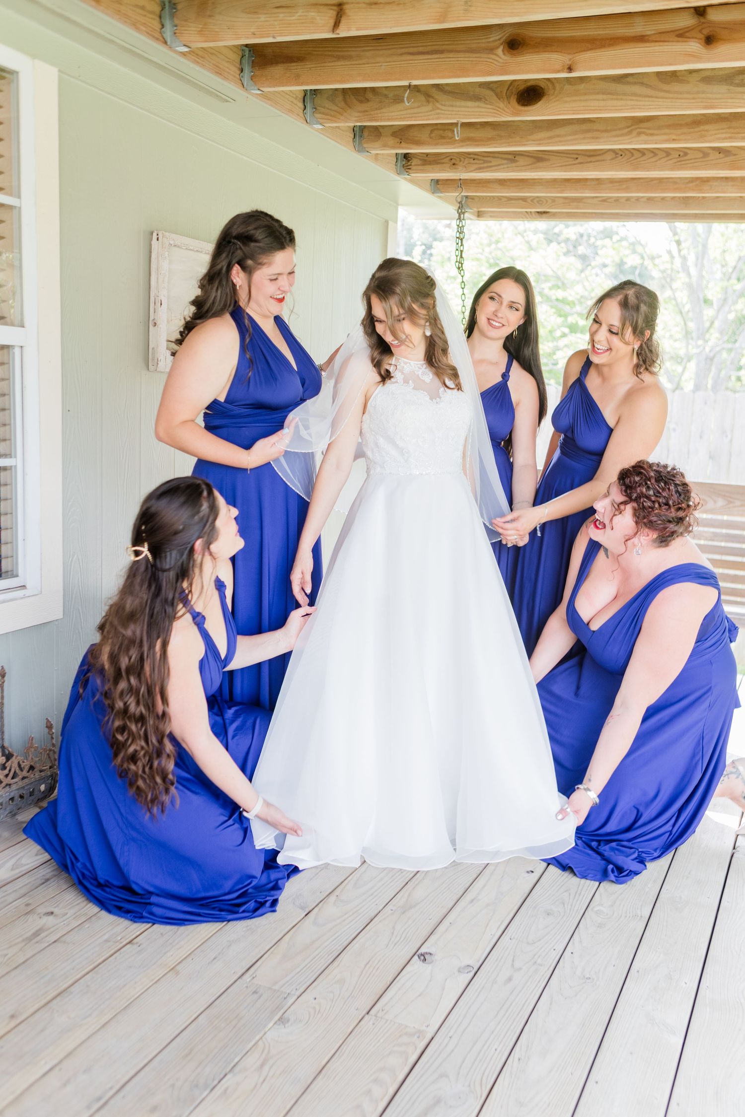 Bridesmaids in blue dresses gathered around white wedding gown on rustic porch.
