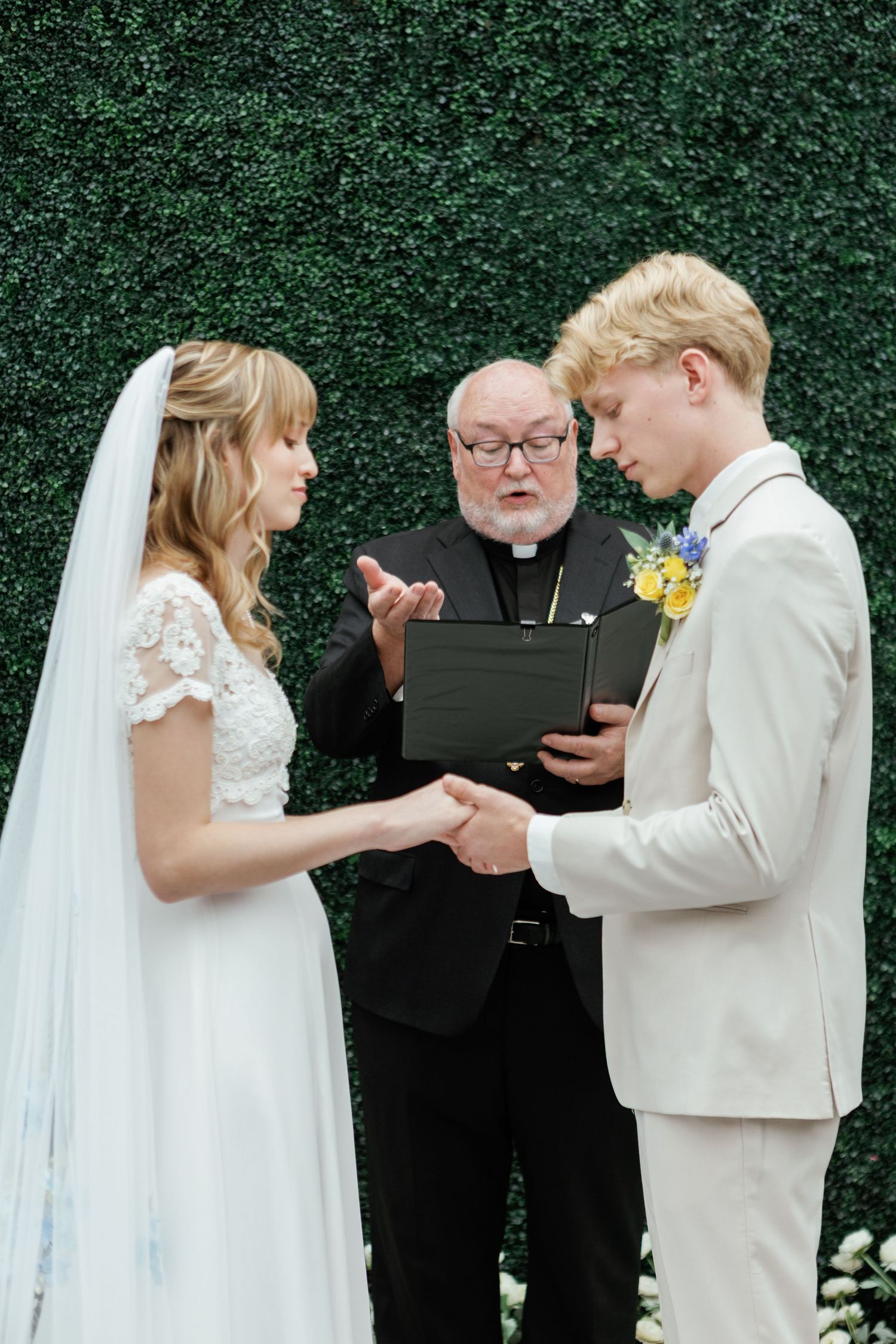 Wedding ceremony scene showing exchange of rings against a green hedge backdrop.