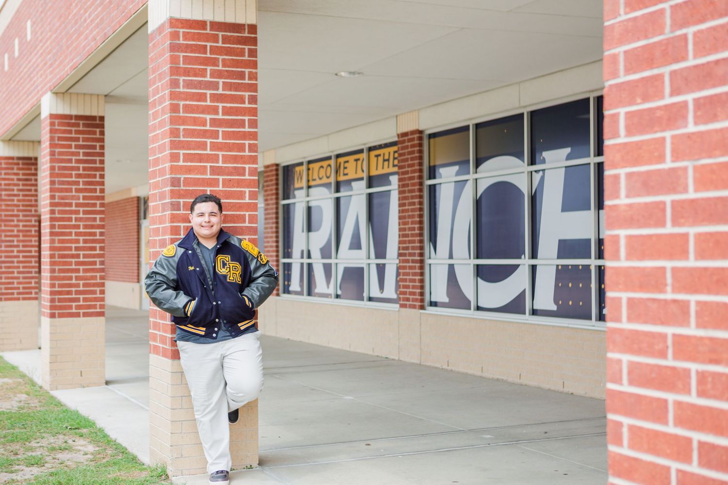 Teen in letterman jacket poses outside brick school building with Ranch sign in window.
