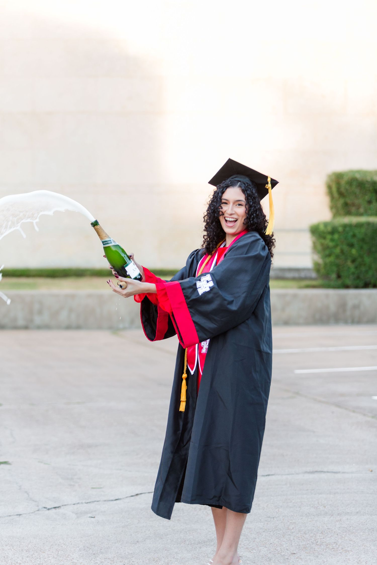 Graduate in cap and gown celebrates with champagne in outdoor setting.