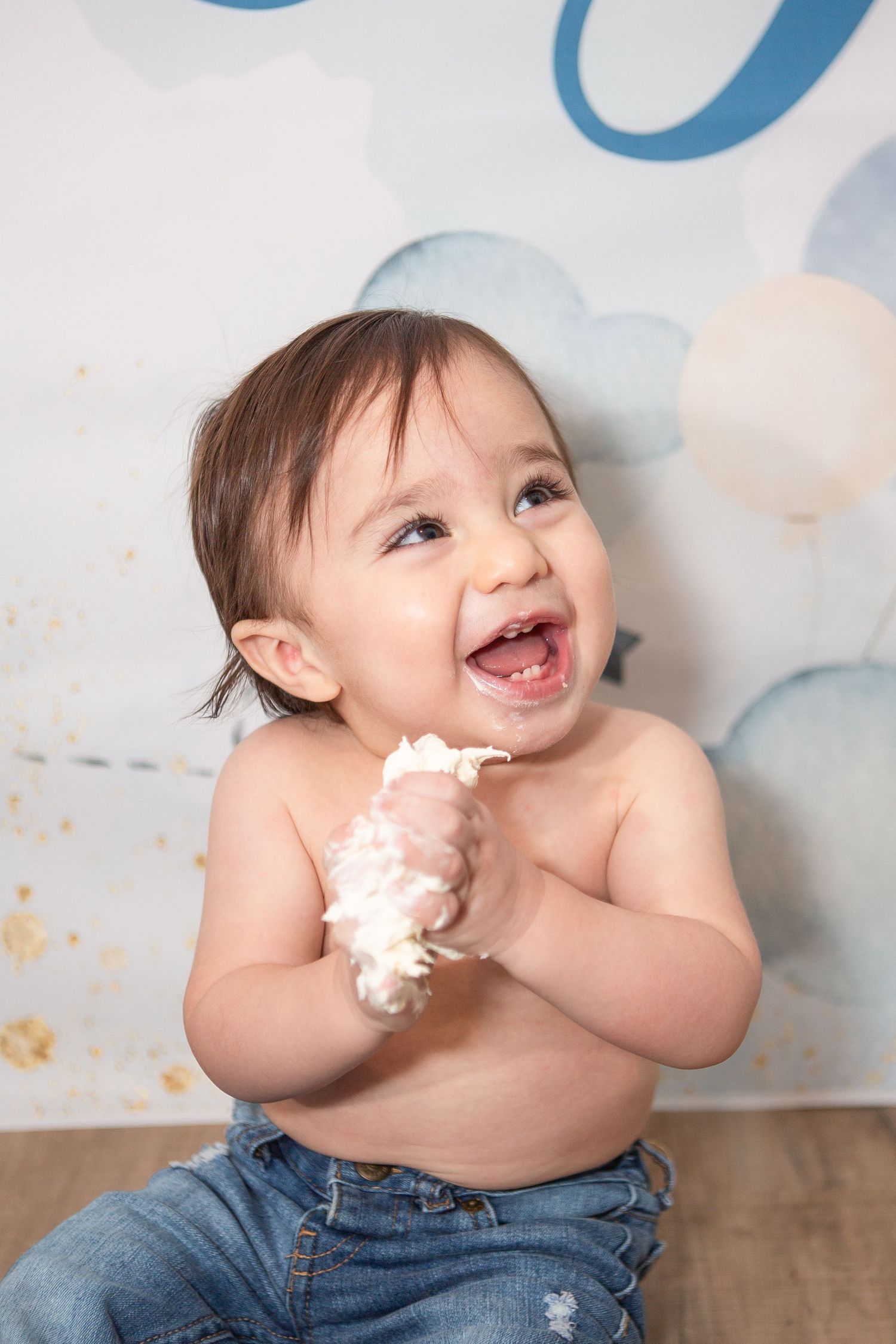 Joyful baby in jeans playing with cake frosting against blue and white backdrop.