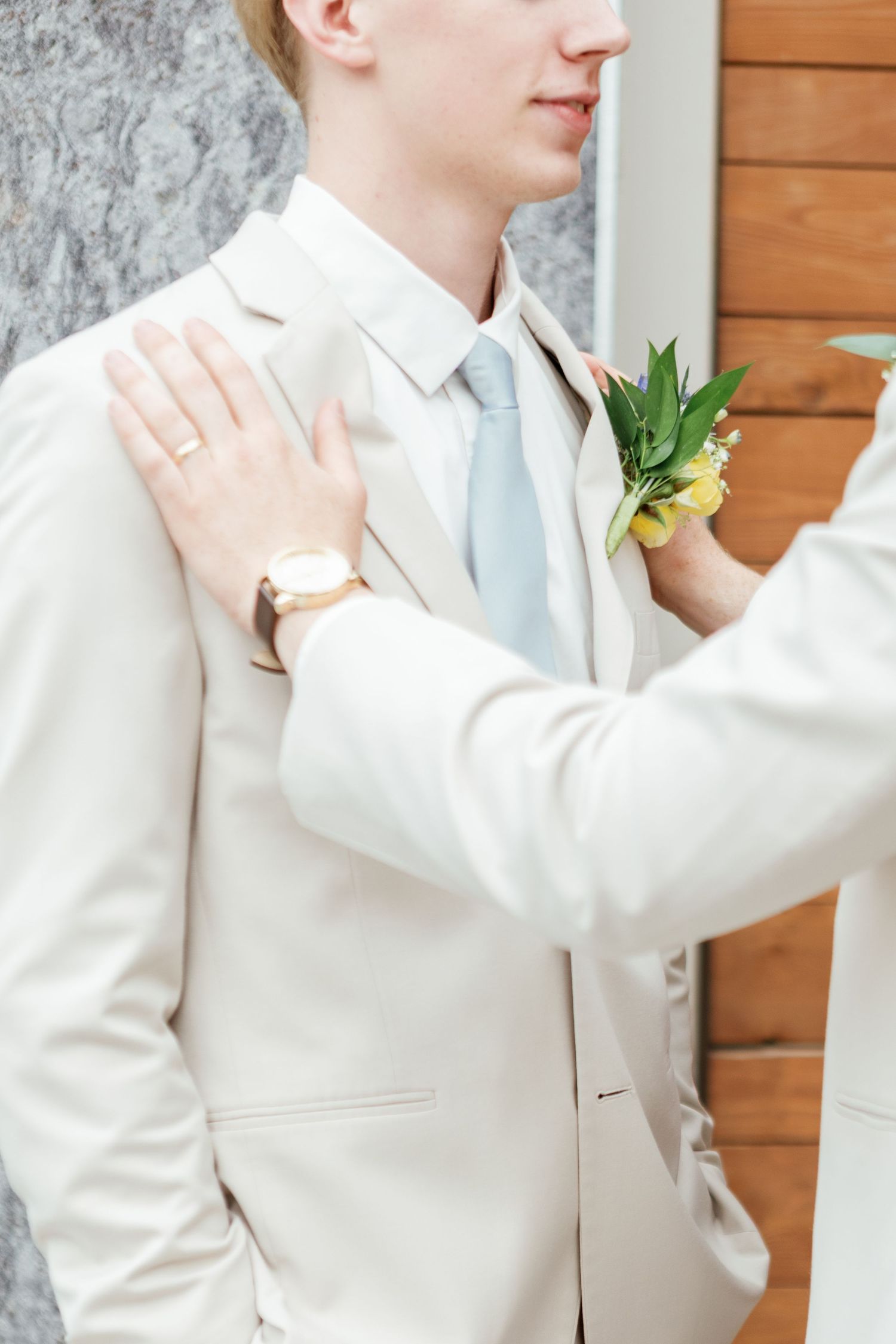 Wedding party member in white suit adjusts boutonniere against wooden wall.