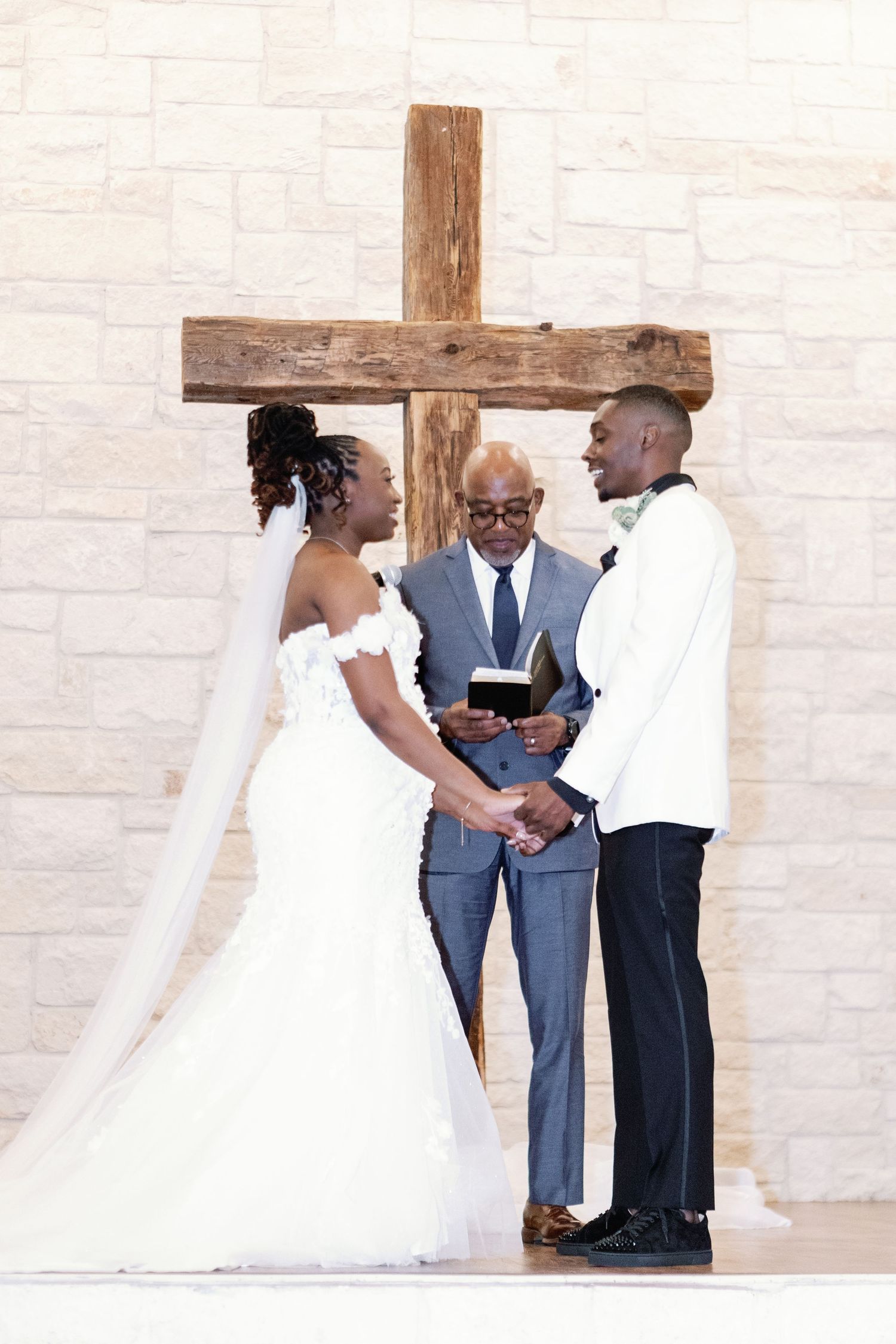 A pair in wedding attire exchanges intimate glances during an outdoor ceremony against a white stone wall.