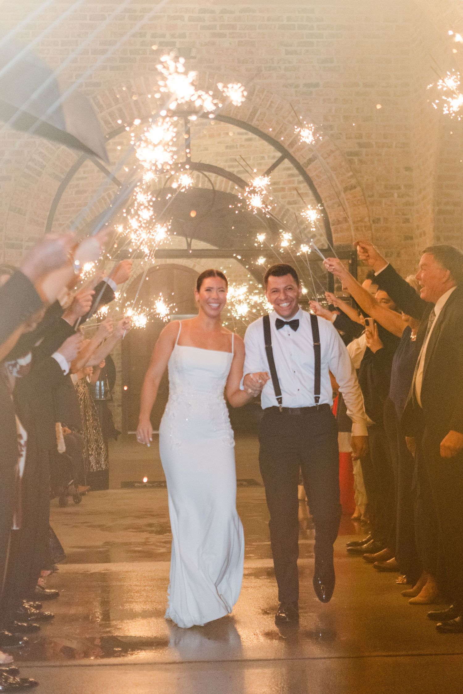 Wedding couple walks under sparklers held by guests in a rustic indoor venue with brick walls.