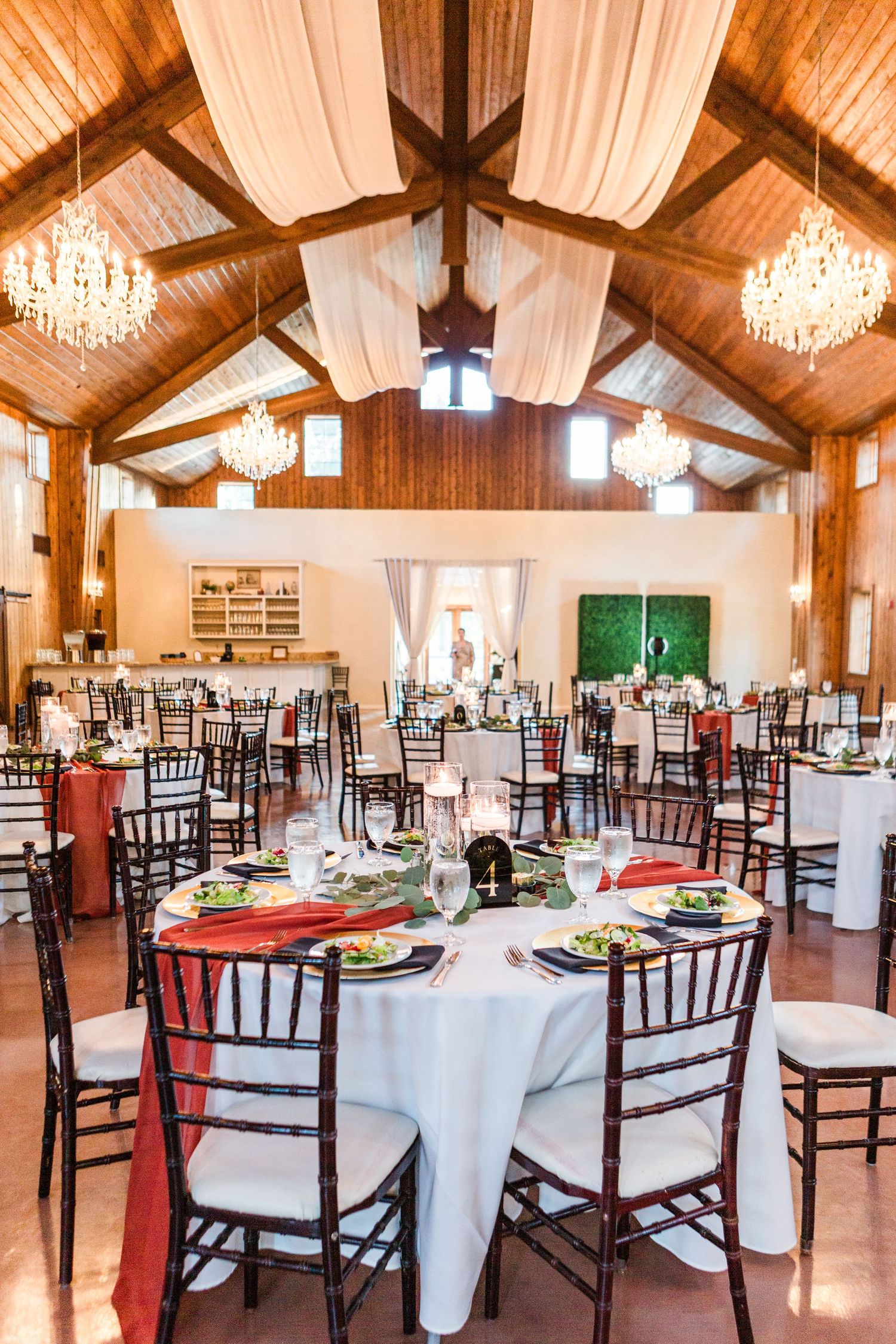 Spacious wedding reception venue with exposed wooden beams, crystal chandeliers and dark chiavari chairs.