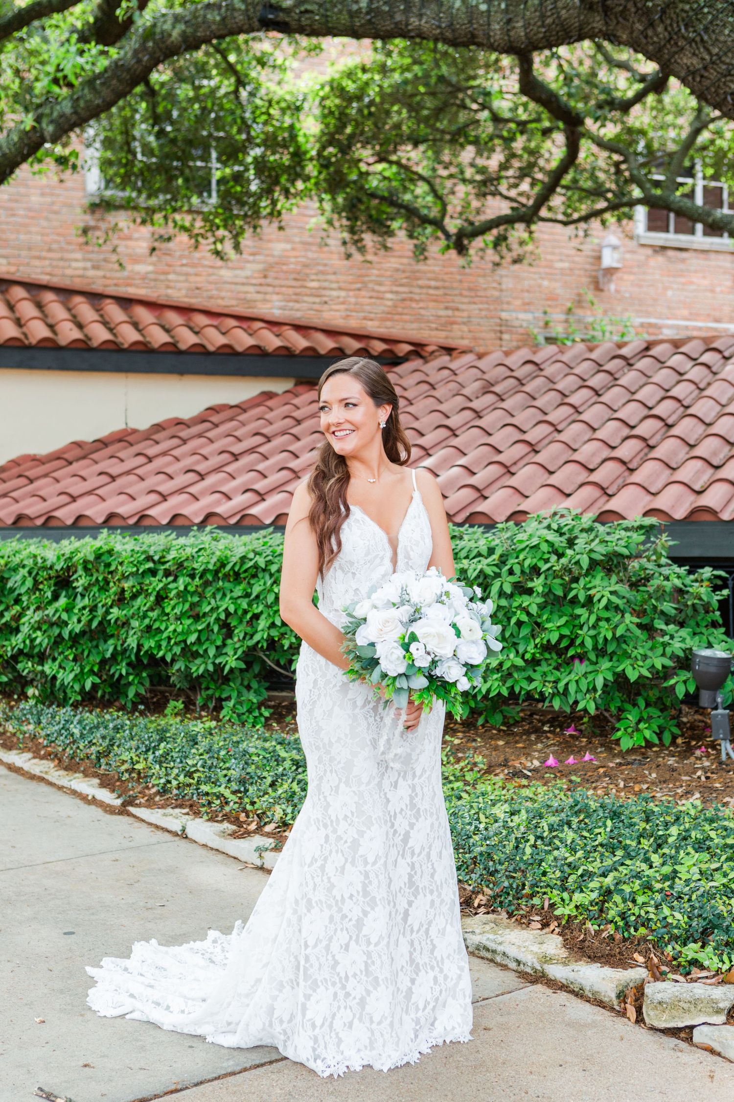 Elegant bride posing with white bouquet in garden setting with terracotta roof.