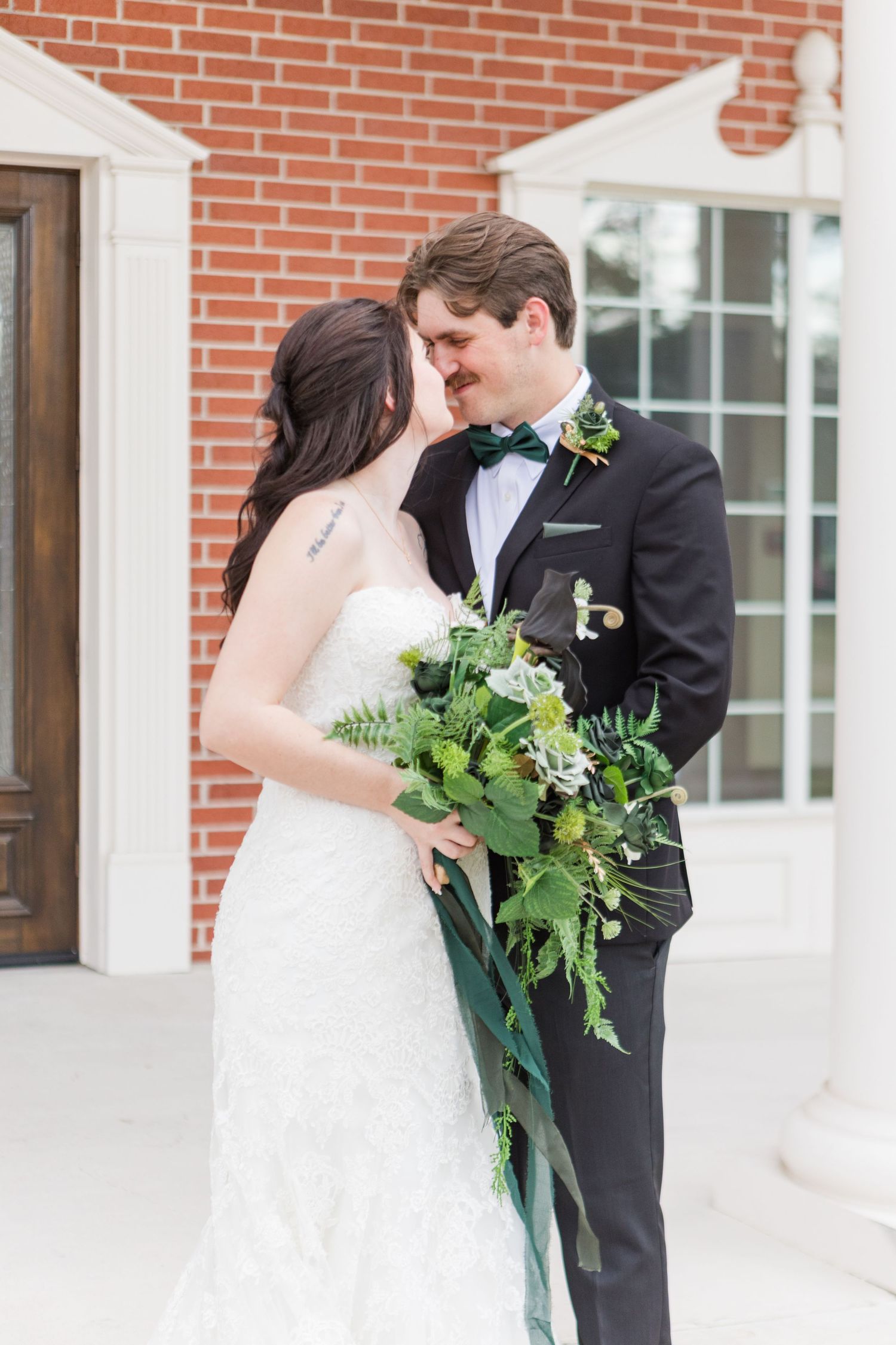 Wedding couple poses against brick wall exterior with white columns and greenery bouquet.