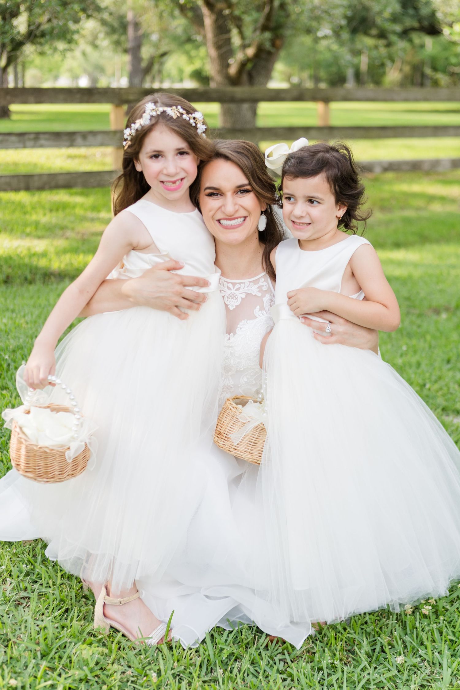 Young attendants in white dresses sitting together on grass at an outdoor wedding.