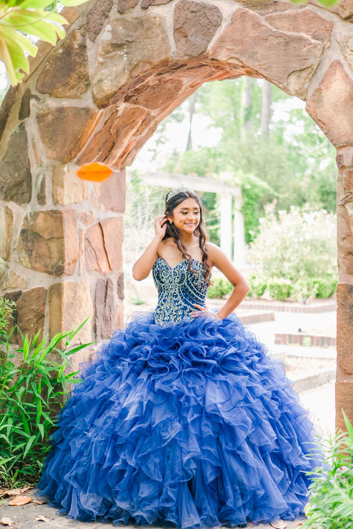 Ornate blue ruffled ball gown showcased under stone archways surrounded by greenery.