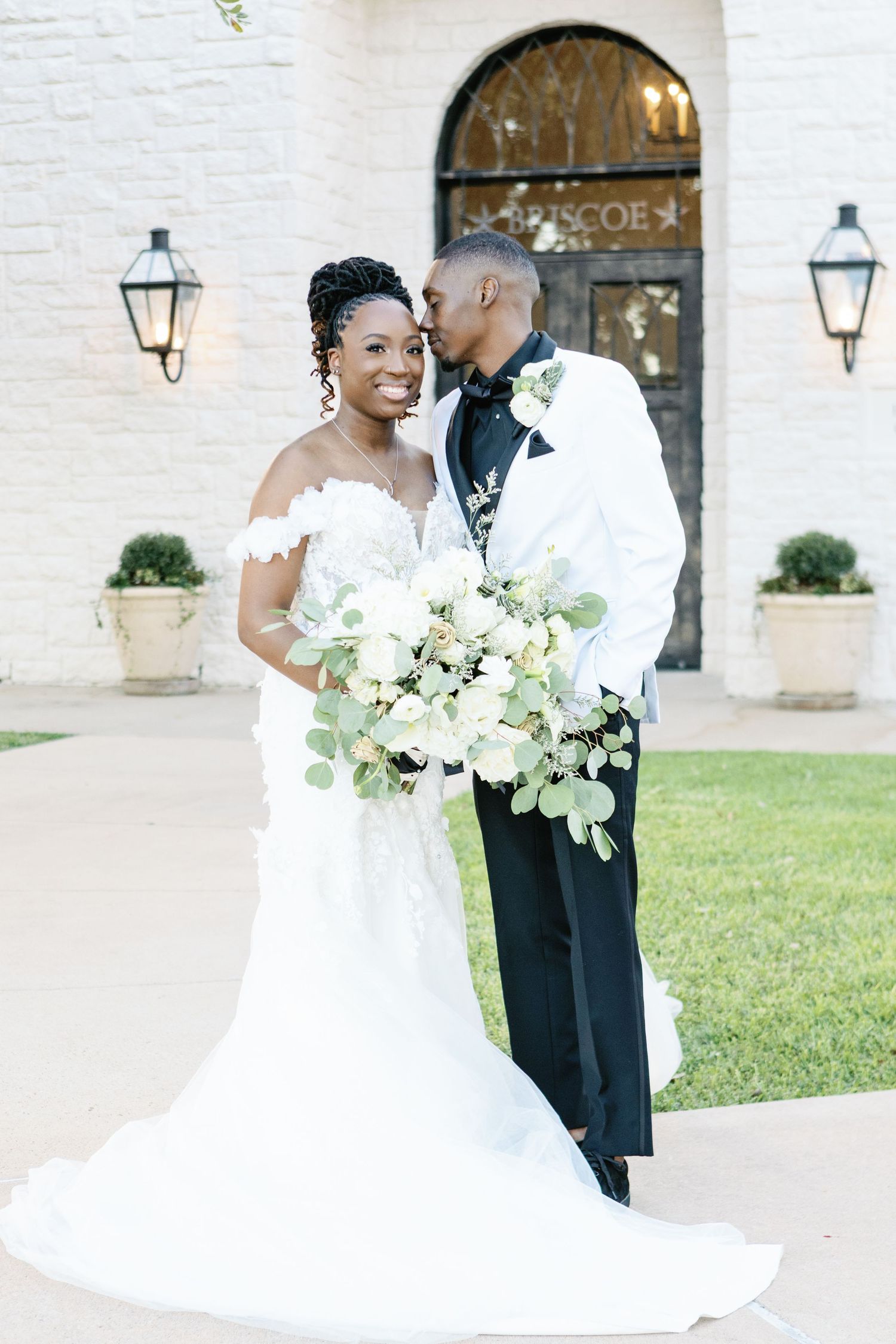 Romantic wedding couple shares intimate moment outdoors against white brick wall with garden lamps and white floral bouquet.