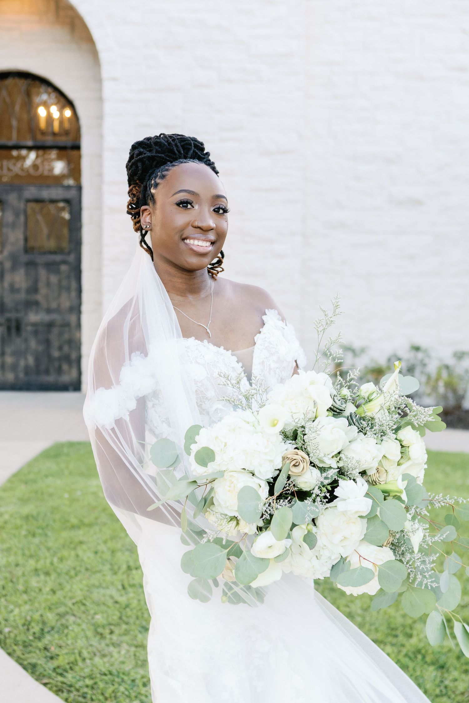 Beautiful bride in white wedding dress holds stunning bouquet of white flowers and eucalyptus outside church venue.