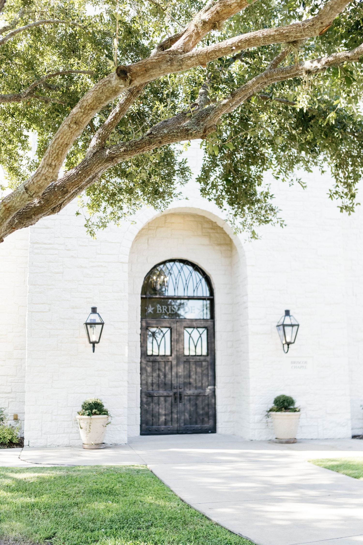 Beautiful white stone chapel exterior with arched doorways and lanterns under sprawling oak trees.