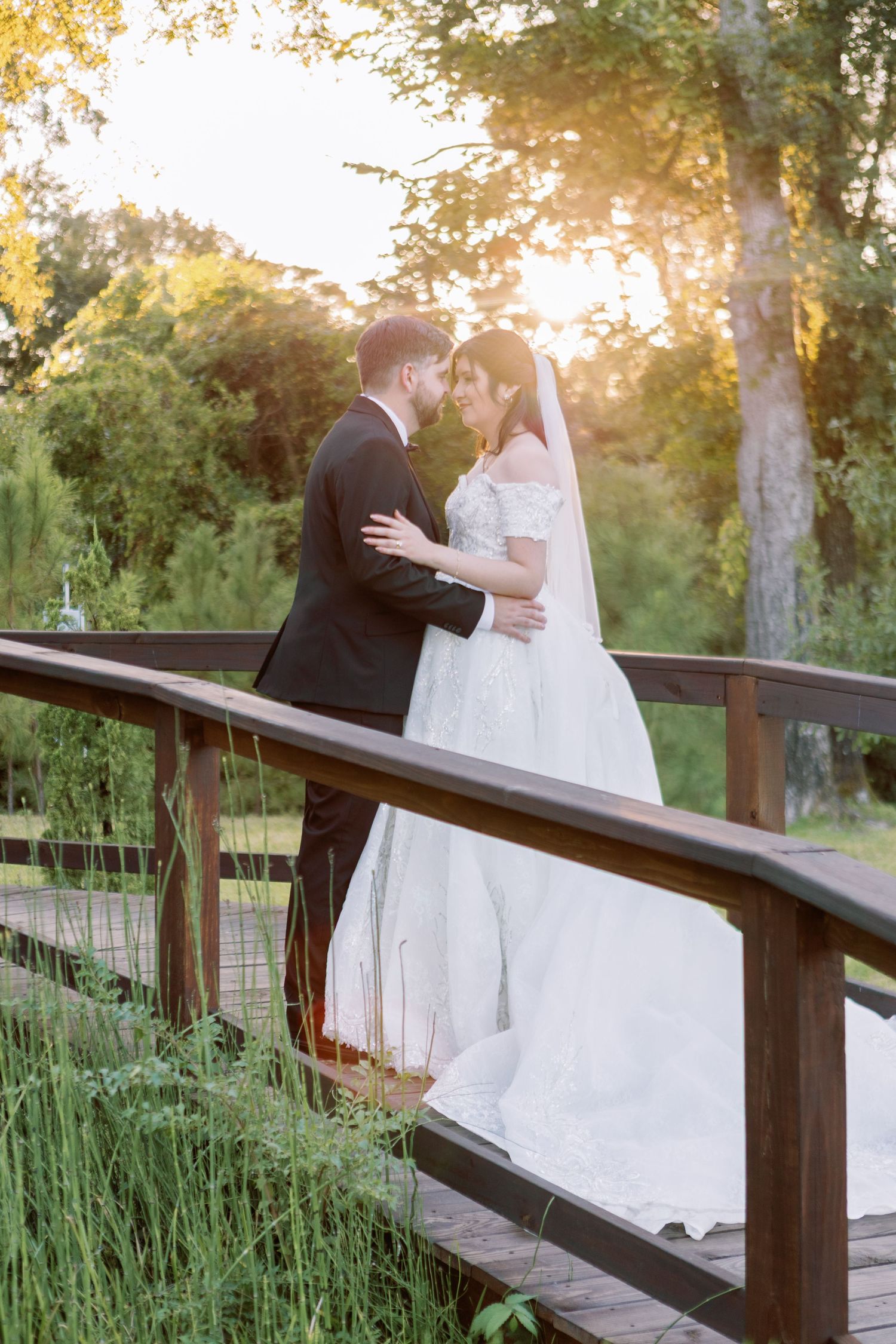 A romantic couple shares an intimate moment at sunset on a wooden bridge surrounded by lush greenery.