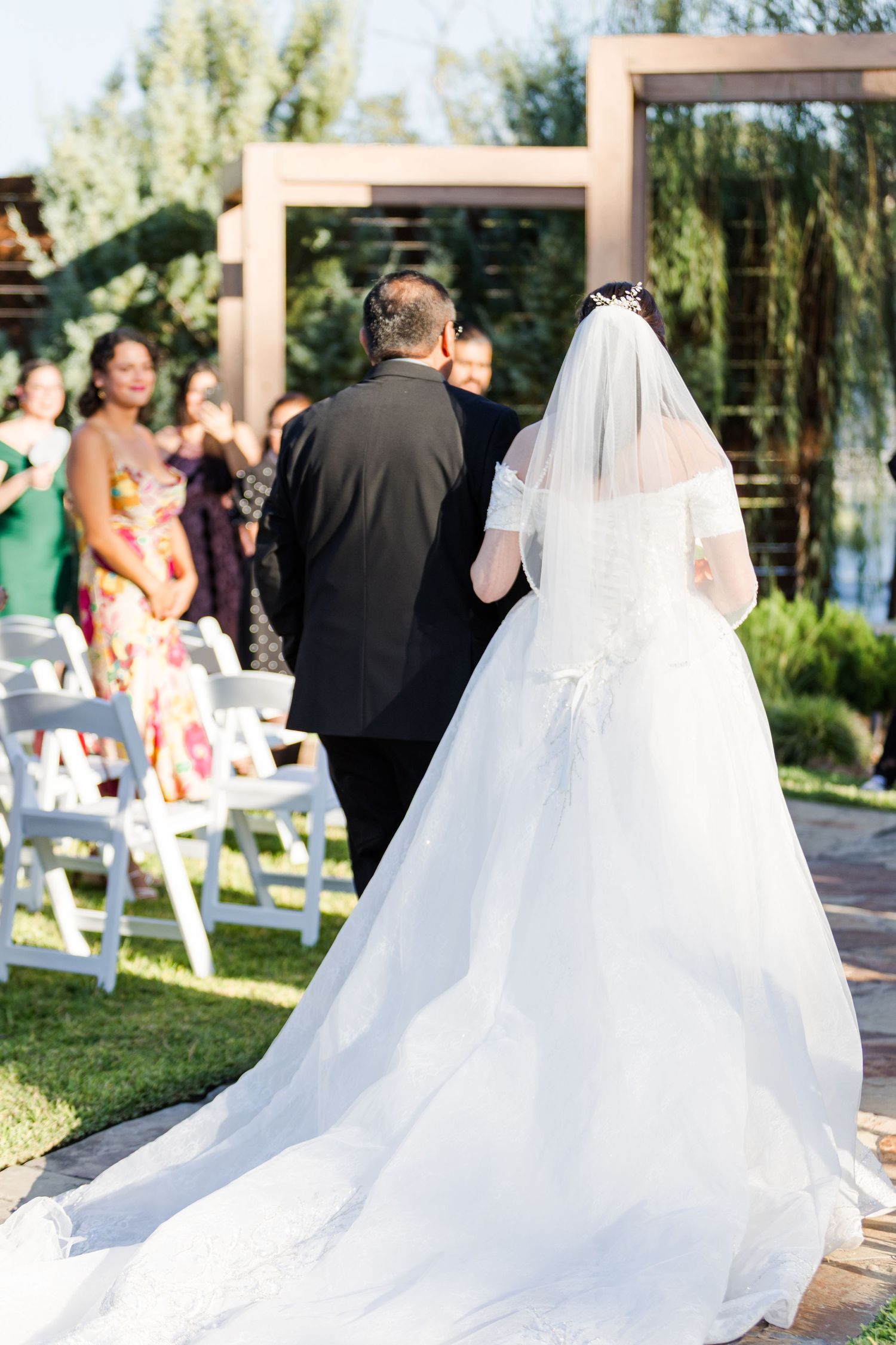 Flowing white wedding dress train trails behind on outdoor ceremony path.