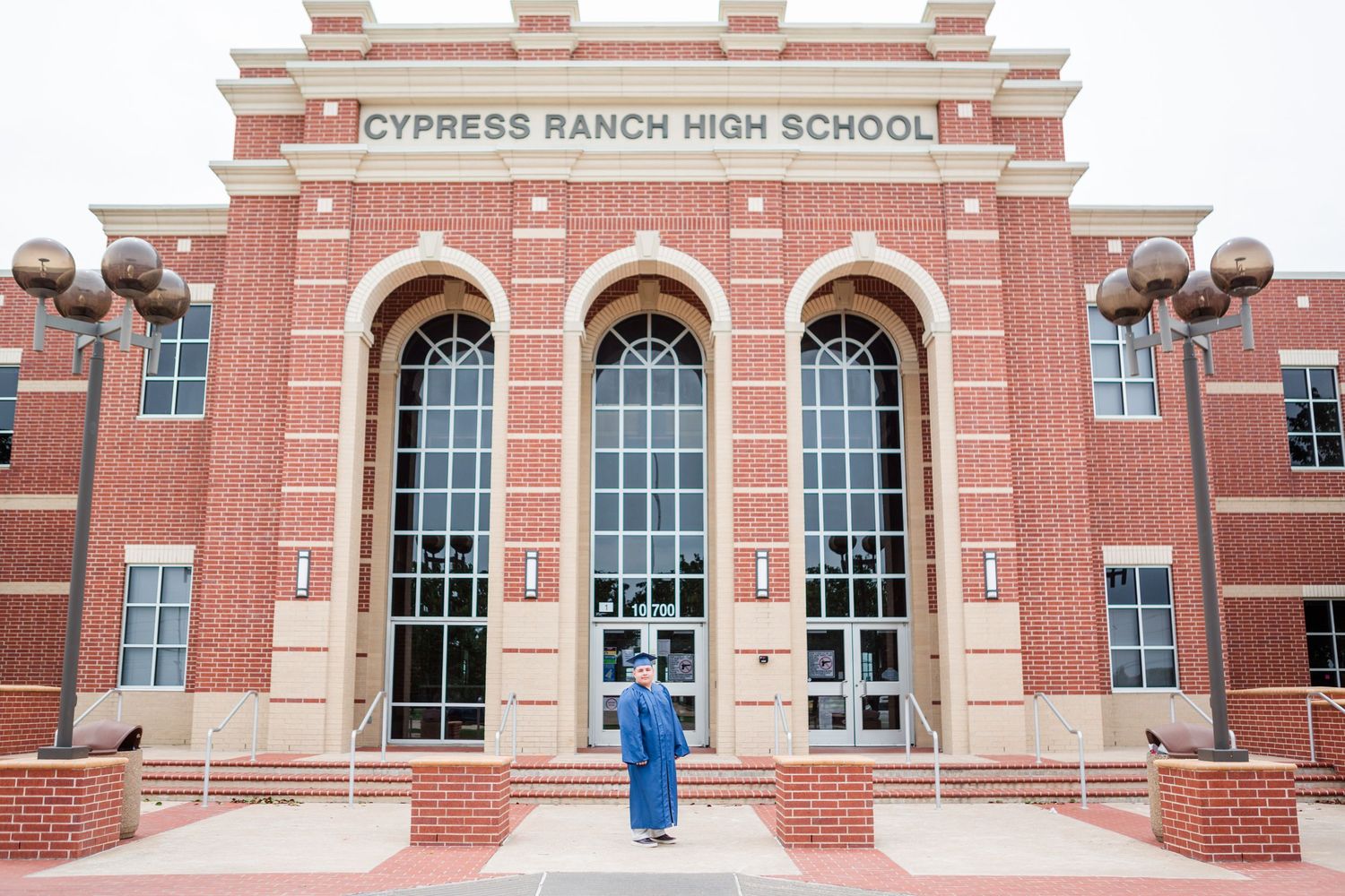 Graduate in cap and gown stands proudly in front of Cypress Ranch High School brick building entrance.