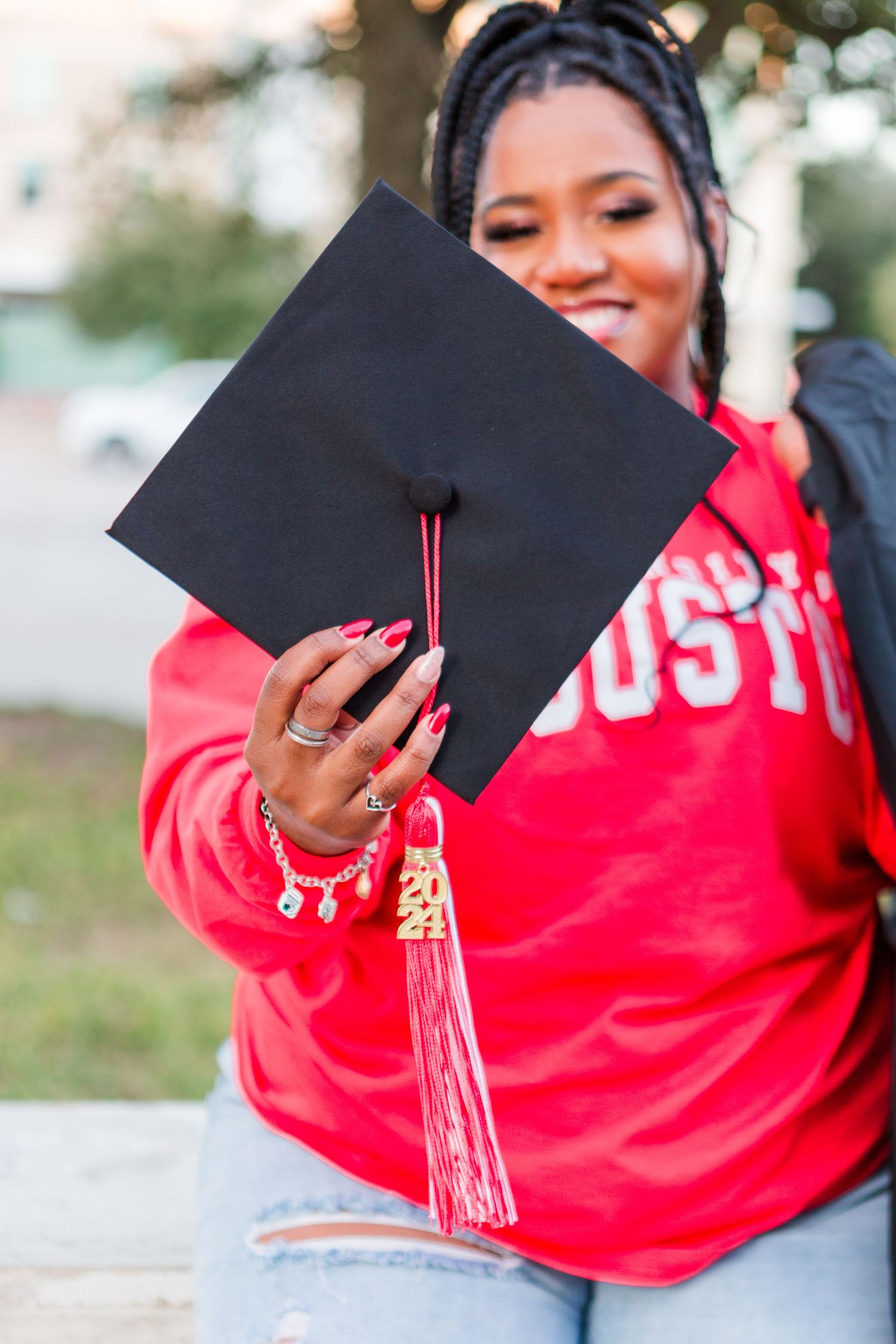 Smiling graduate in red Houston sweatshirt holding black graduation cap with red tassel outdoors.