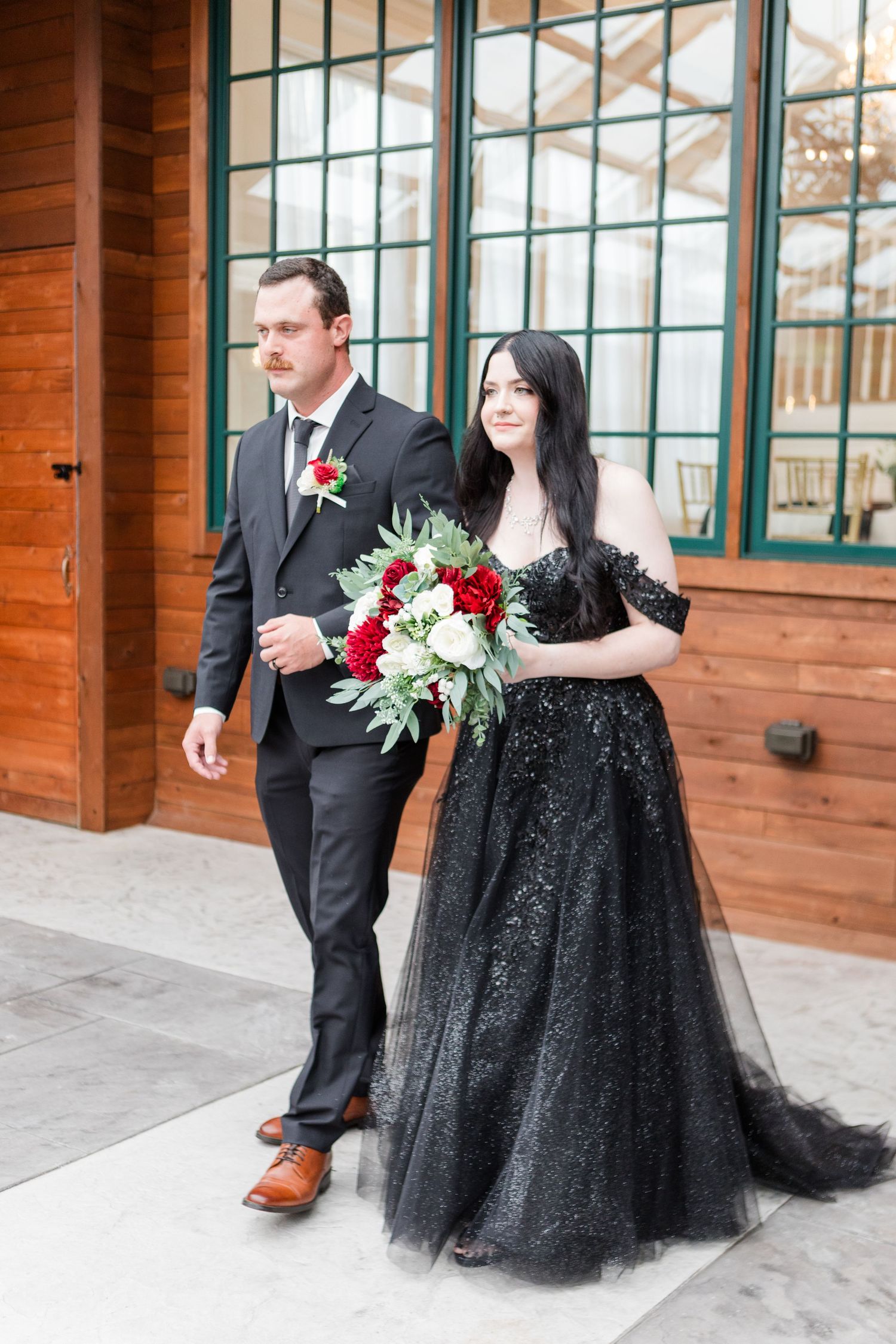 Wedding couple in black attire pose outside rustic wood building with large windows.