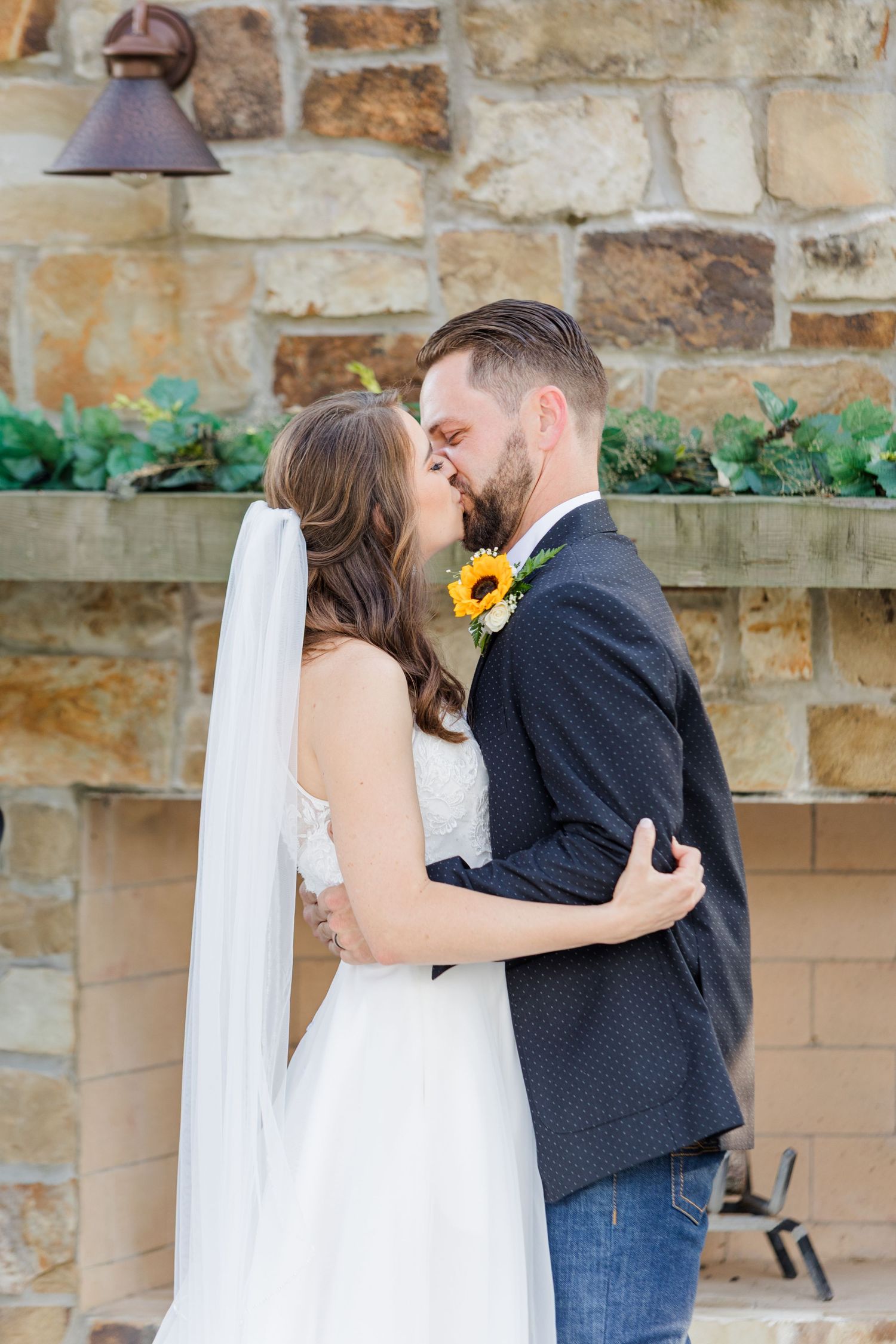 Romantic wedding moment captured against rustic stone wall with sunflowers.