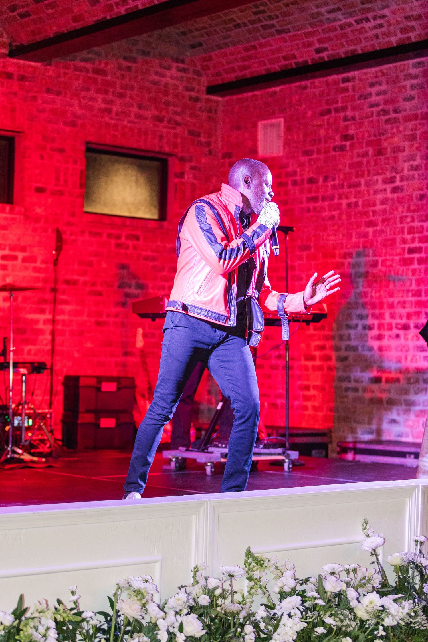 Performer in blue jeans and vest dances energetically on stage with red brick wall backdrop and dramatic lighting.