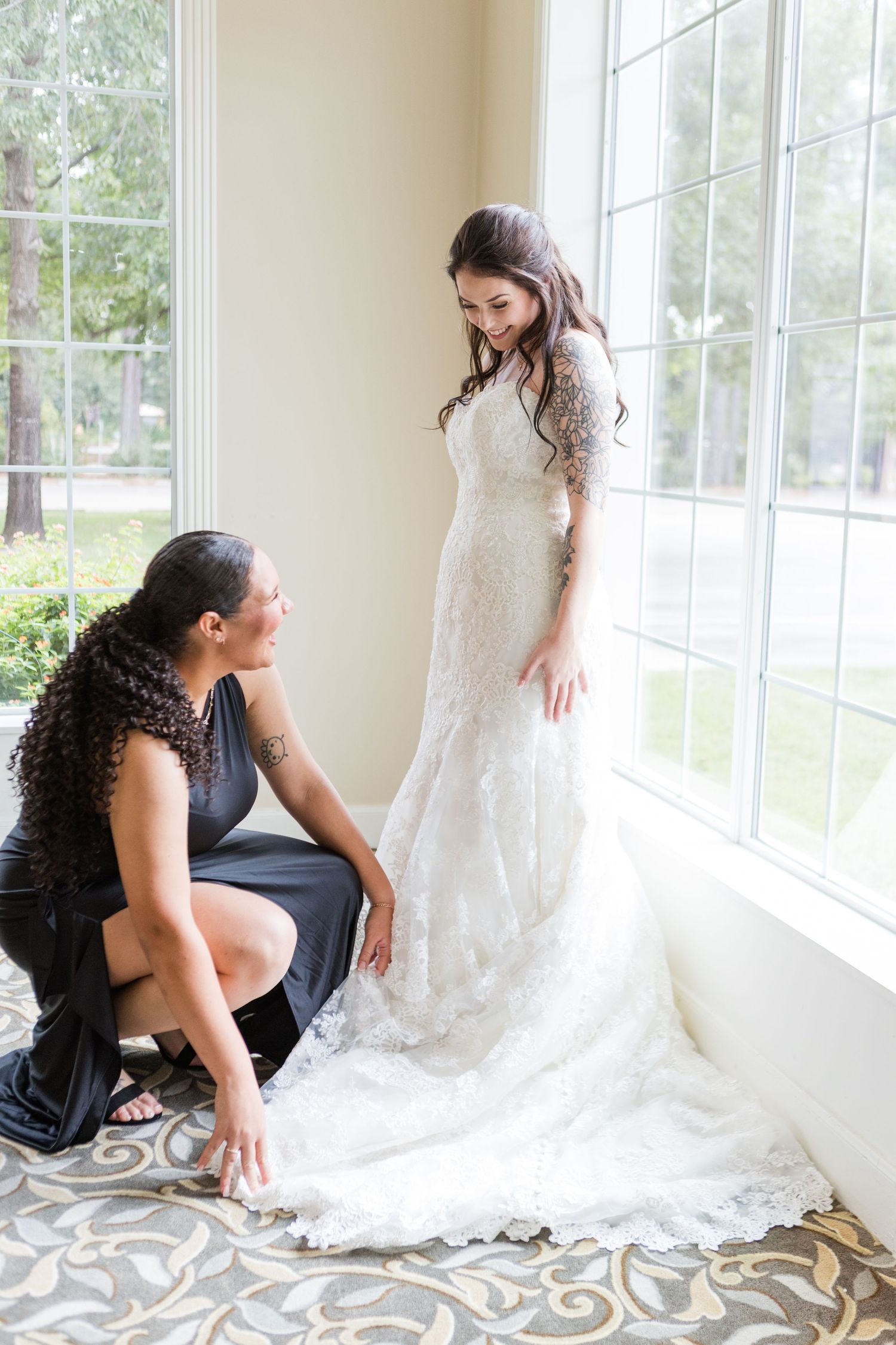 A flowing white wedding dress train is arranged on a patterned carpet near bright windows.