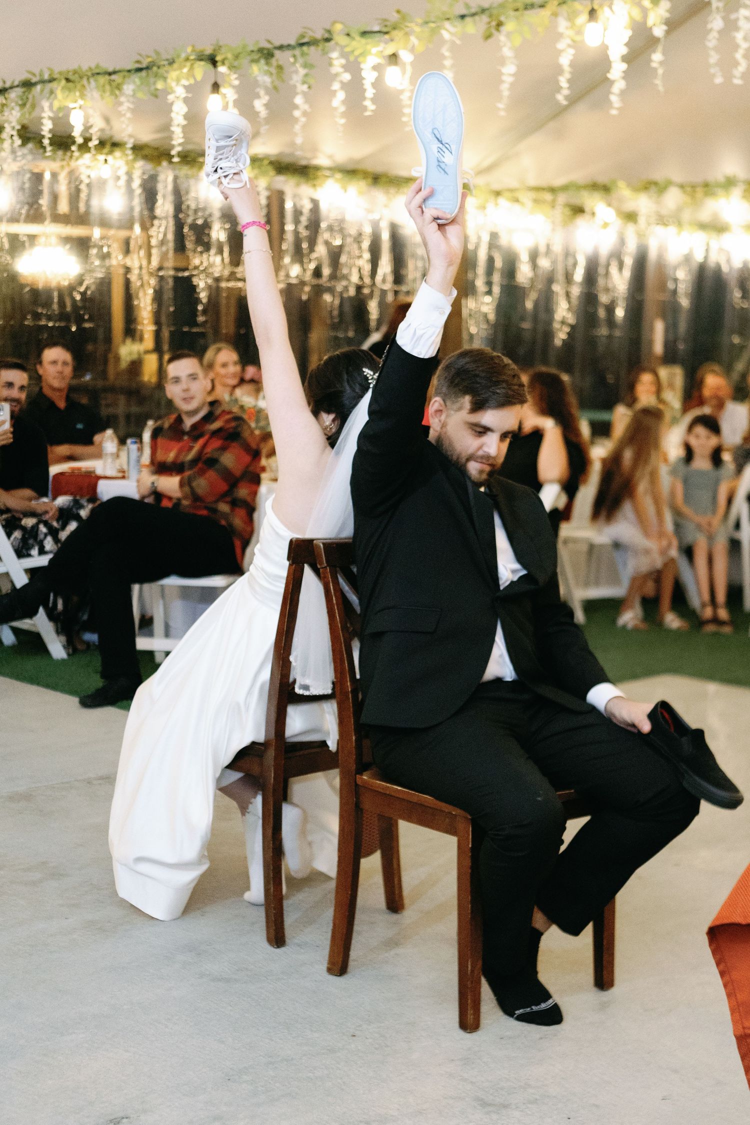 Wedding reception game with couple sitting on wooden chairs under string lights.