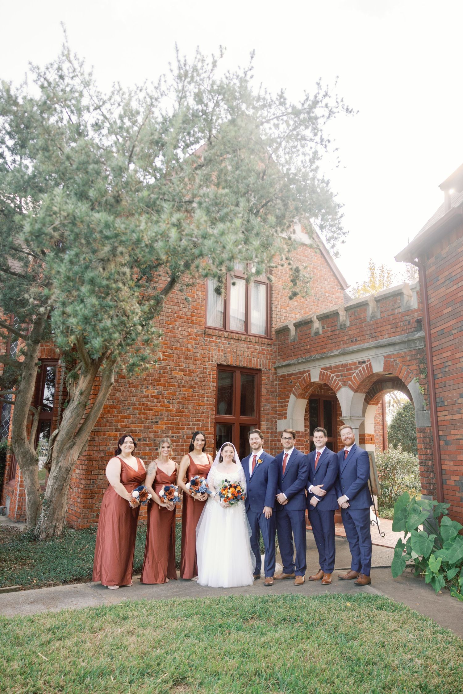 Wedding party in navy blue and burgundy attire poses together in front of brick church with olive trees.