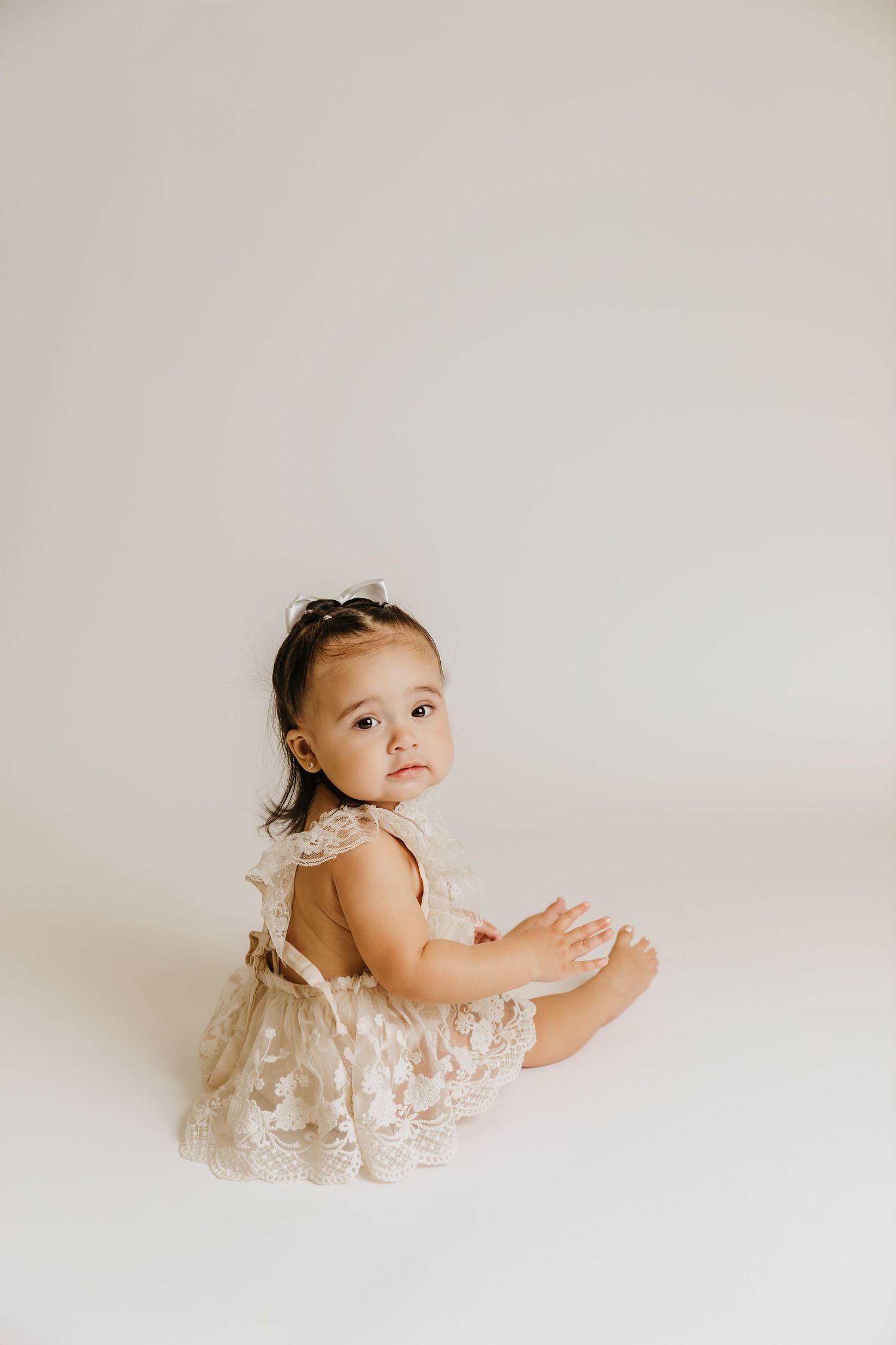 One-year-old girl sit on the floor in-studio looking back over her shoulder during first birthday photoshoot with Tami Harris Photography