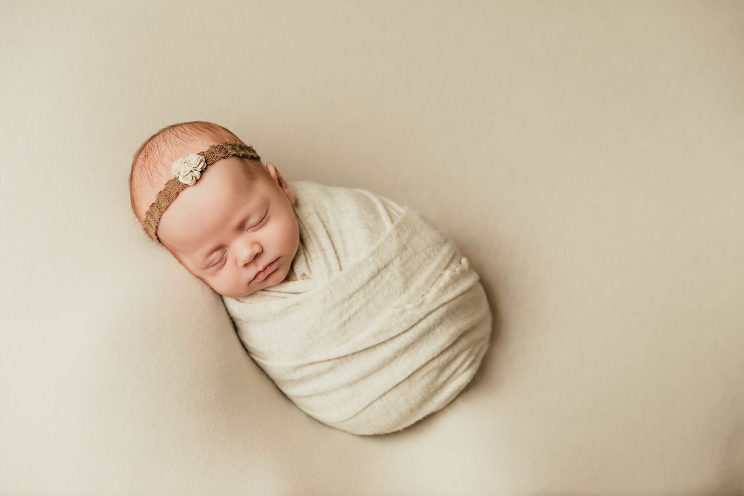 Newborn baby girl sleeping peacefully while swaddled in cream wrap and brown headband during photoshoot with Tami Harris Photography